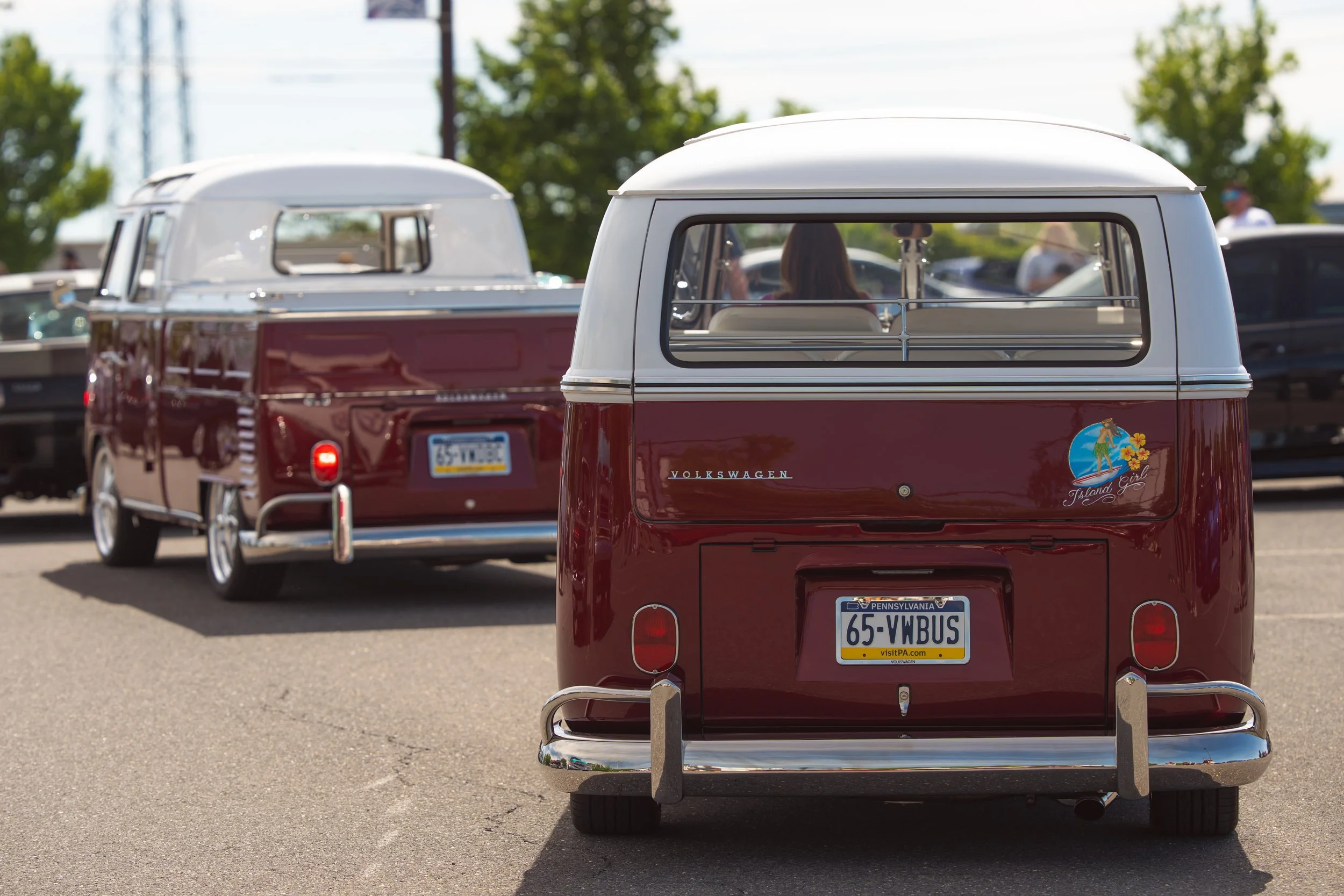 Two vintage Volkswagen Type 2 vans parked in an outdoor lot. The vans are maroon with white roofs and have Pennsylvania license plates. The back of the vans features a sticker or decal, one with an island scene and the words "Island Girl." The backgr