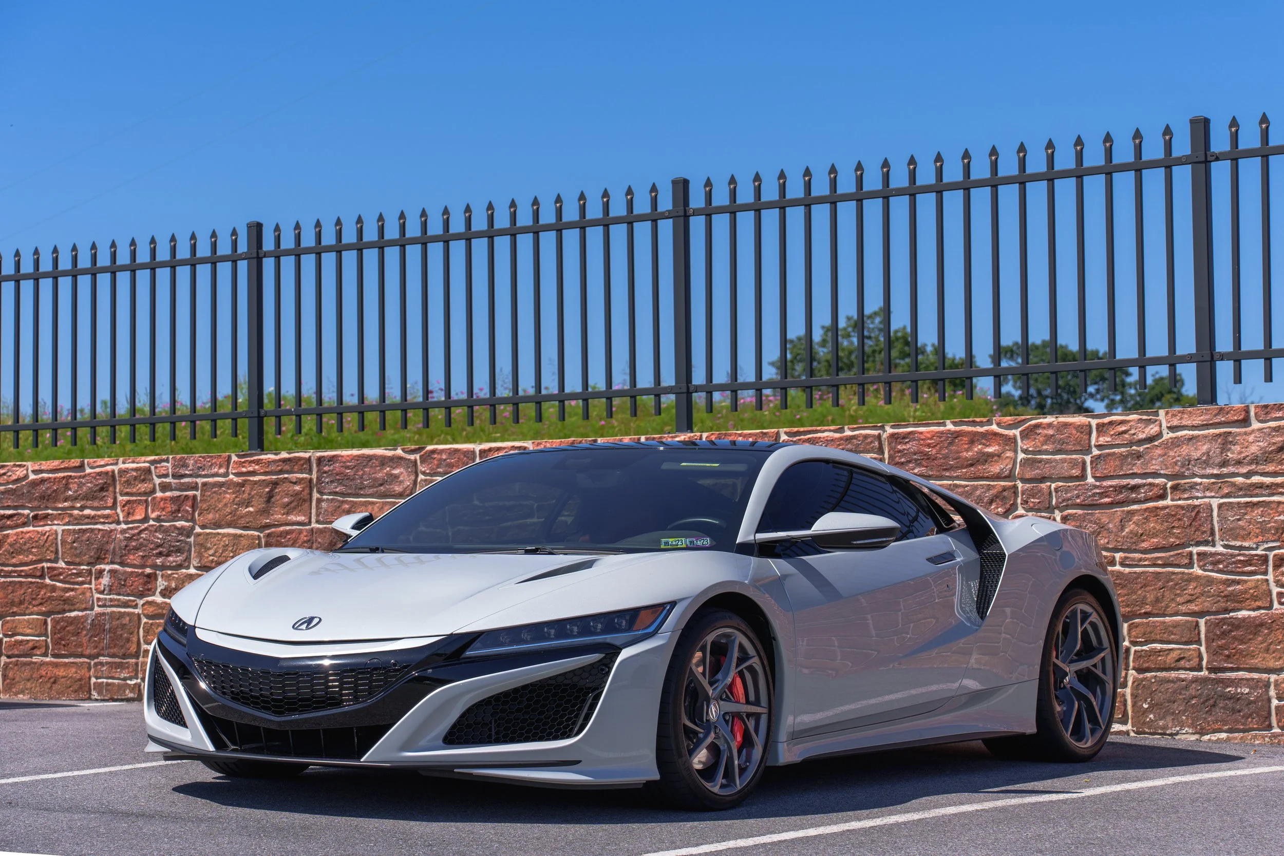 A white sports car parked in a parking lot in front of a brick wall, with a black metal fence above the wall and blue sky in the background.