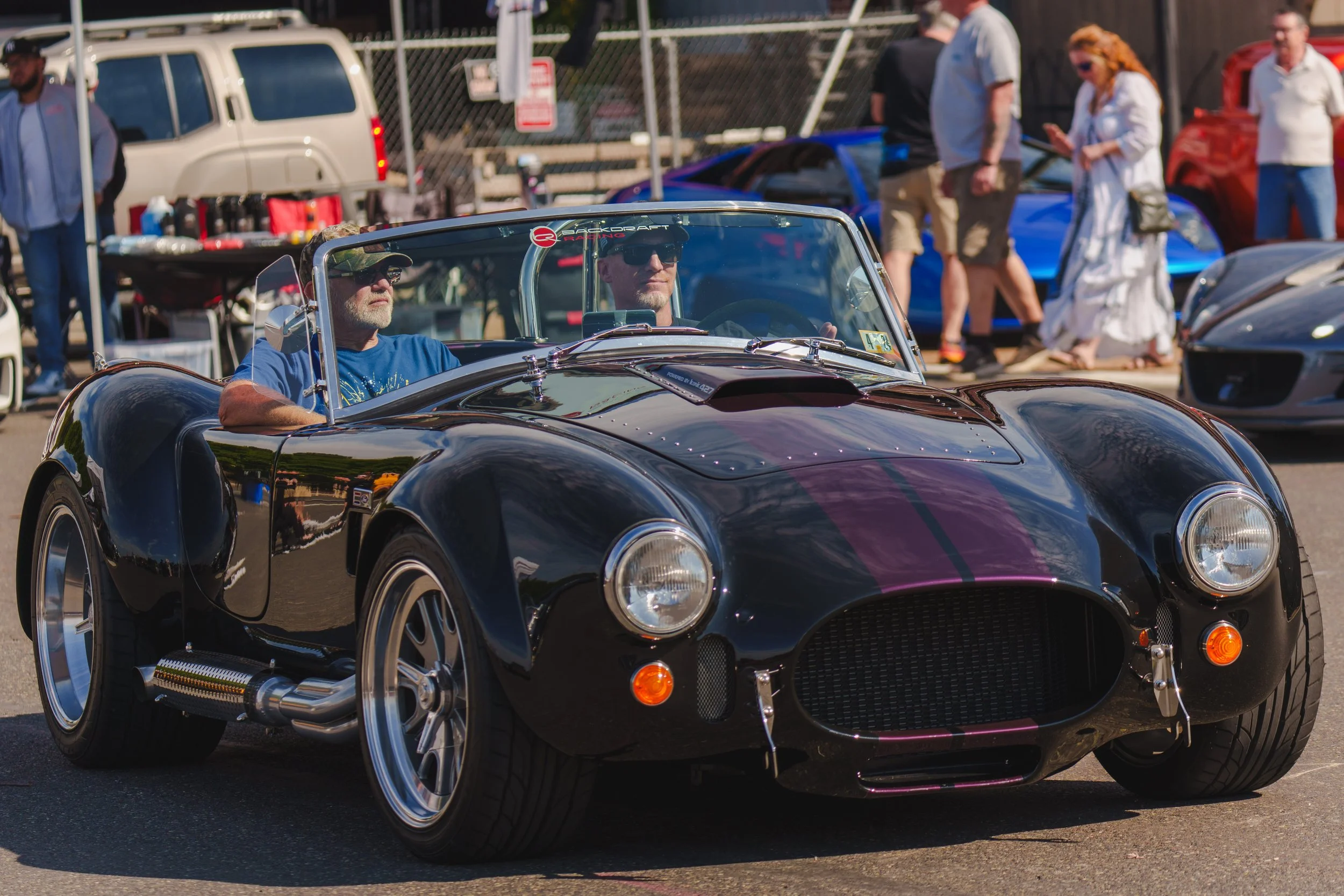 A black vintage sports car with purple racing stripes on display at an outdoor car show. Two men wearing sunglasses sit inside the car. People are walking and observing other cars in the background.