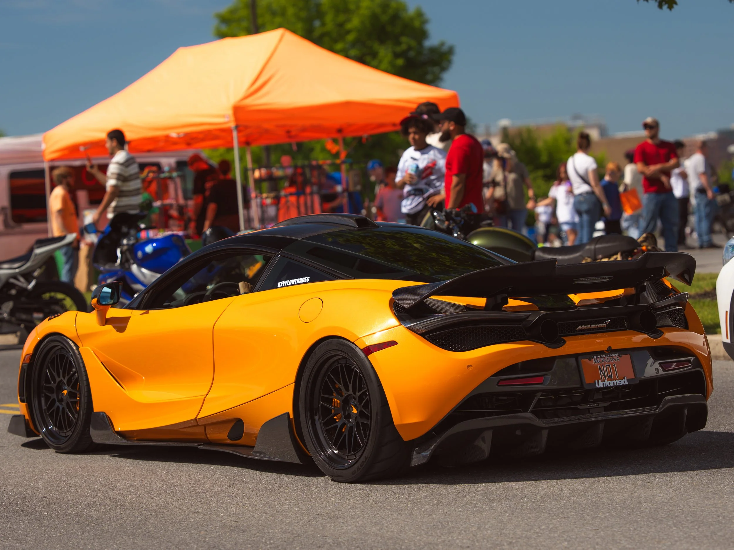 A bright orange sports car parked on a street during an outdoor event, with a crowd of people and vendor tents in the background.