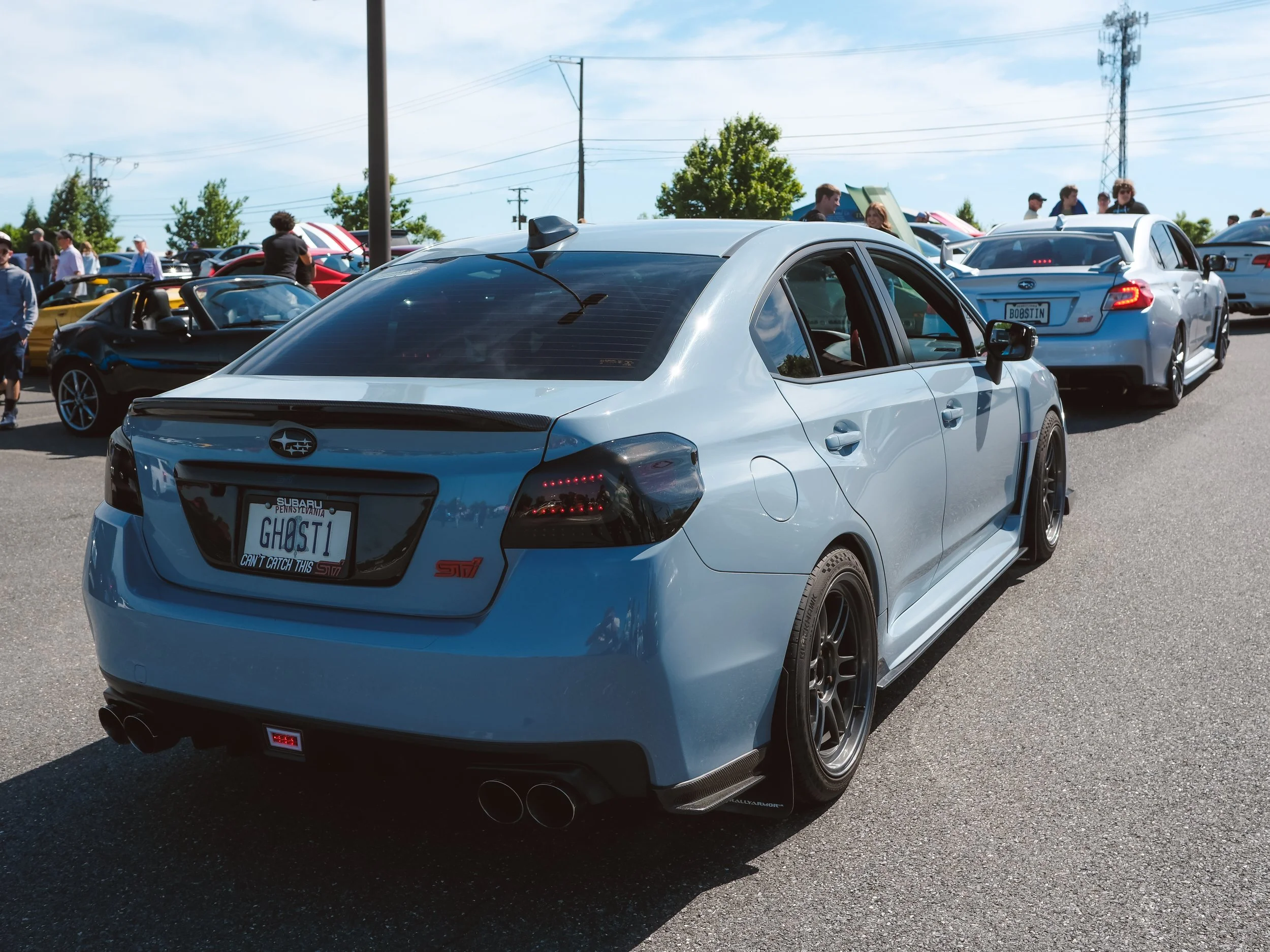 A silver Subaru WRX with a custom license plate reading 'GHOST1' and a decal that says 'Can't Catch This' at a car meet, with other cars and people in the background on a sunny day.