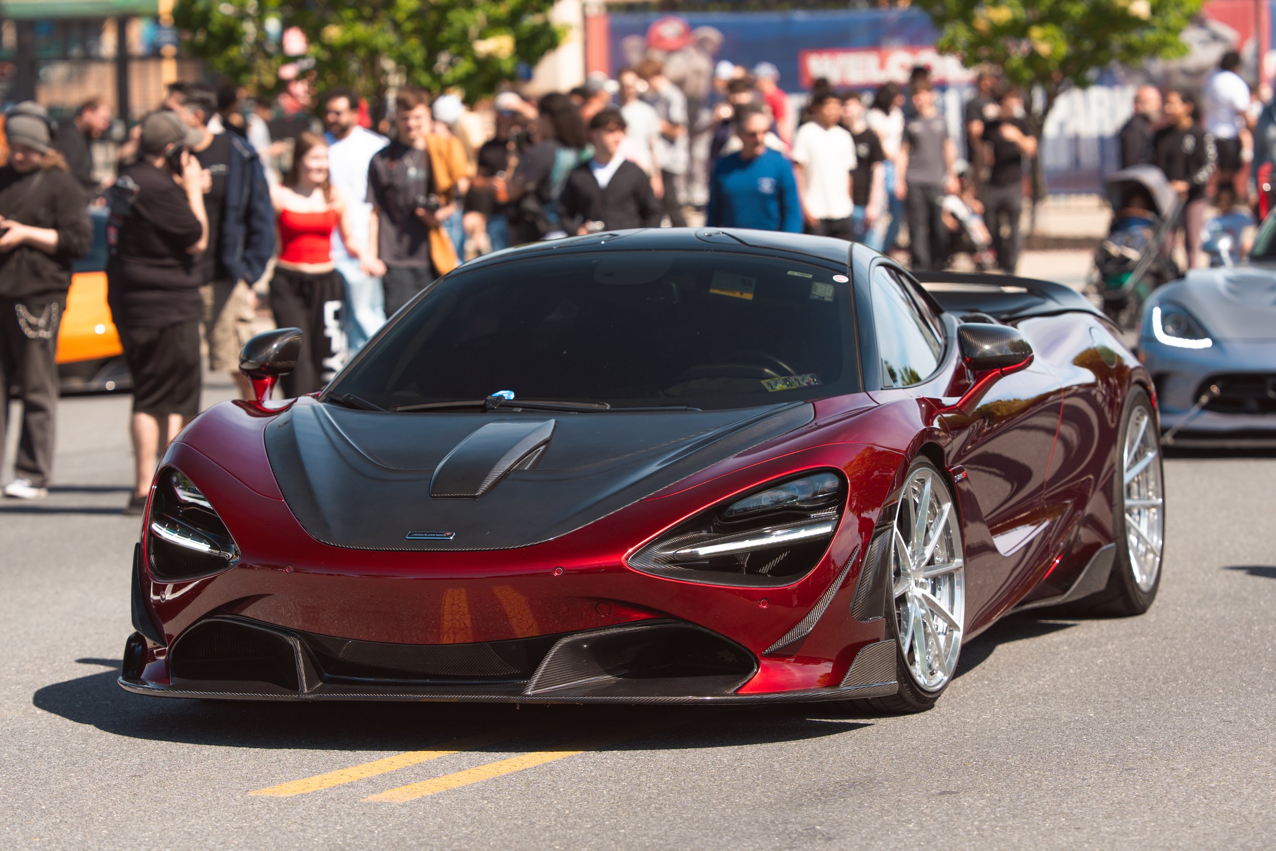 Close-up of a red sports car with sleek design, parked on a street with a crowd of people and other vehicles in the background.