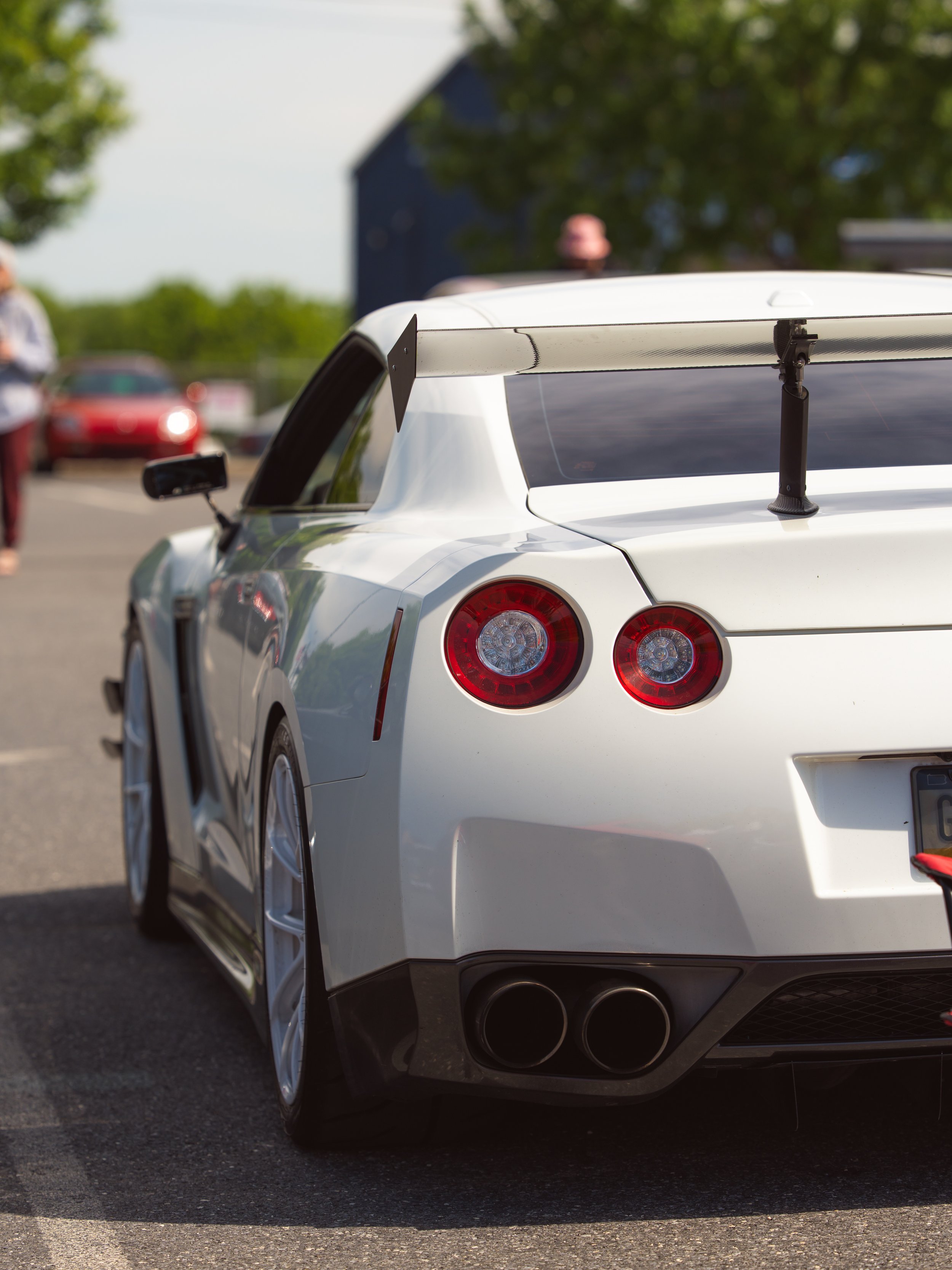 The rear part of a white sports car with a large rear wing, round taillights, and quad exhaust pipes parked outdoors on a sunny day.