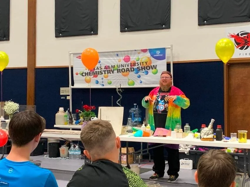 A man in a colorful tie-dye jacket demonstrating science experiments at a university chemistry roadshow event. Students watch in the foreground. The background has a banner, balloons, and science equipment on tables.
