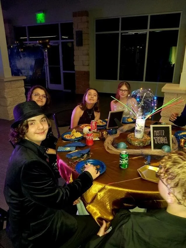 Group of five children sitting around a decorated birthday party table with plates of food, drinks, a birthday cake with sparklers, and a sign reading 'Master Yoda' in a dimly lit room.