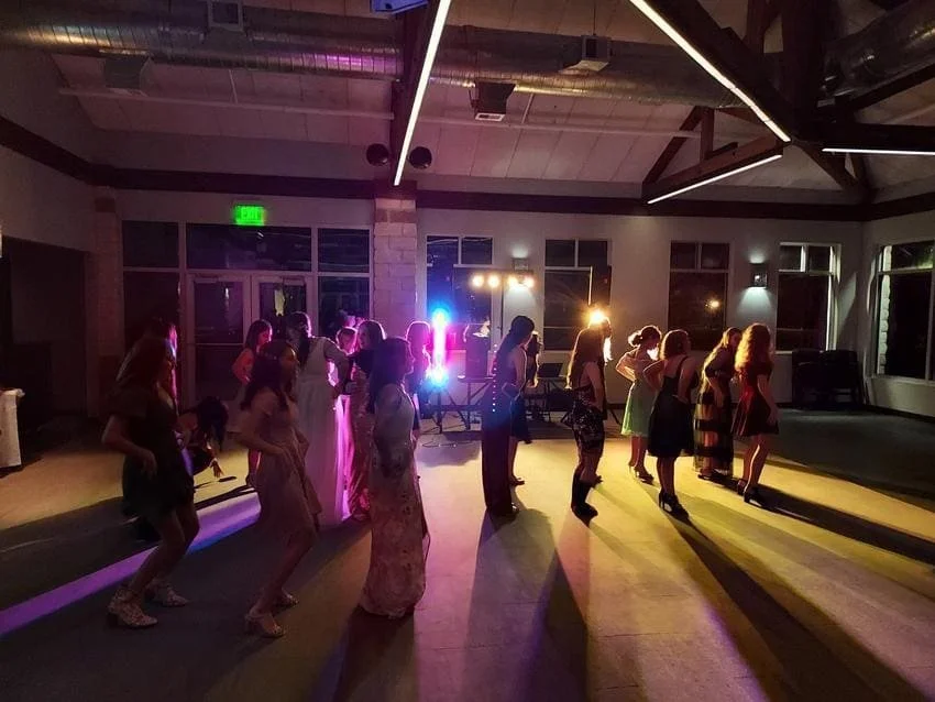 People dancing at a party or celebration in a dimly lit venue with colorful lighting and large windows.