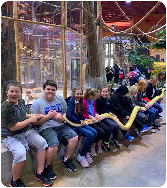Group of children sitting in a row at an indoor play area holding a large yellow snake.