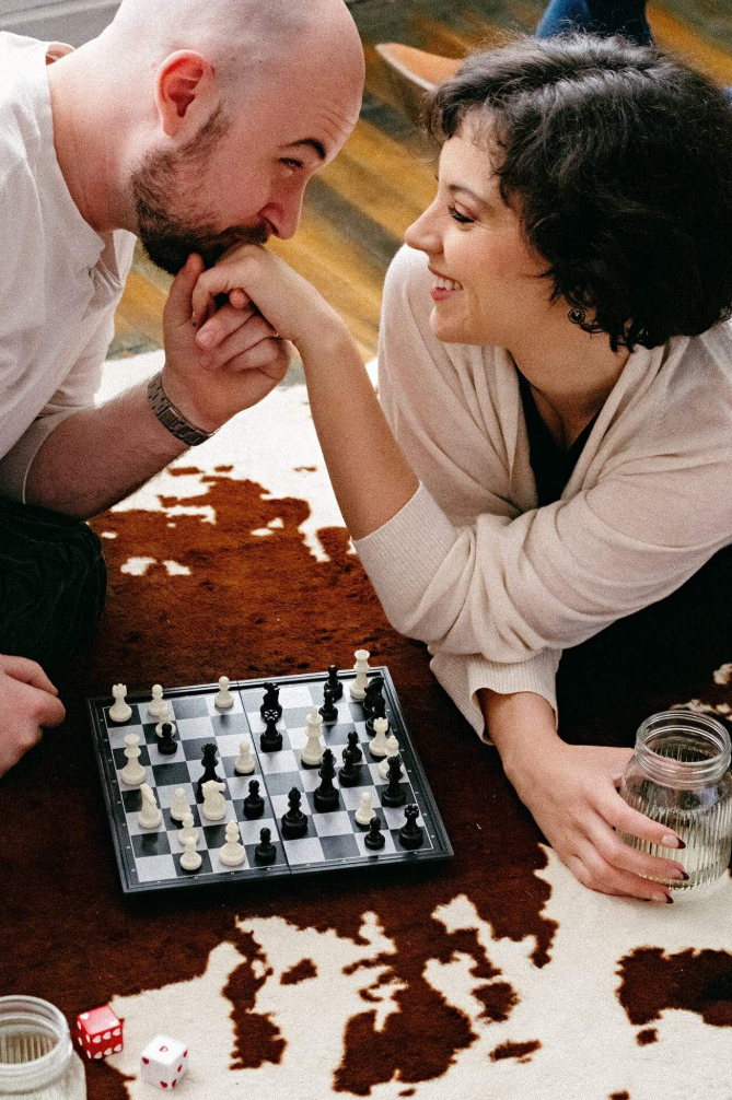A man and woman are sitting on the floor playing chess, smiling, with the man kissing the woman's hand. There are jars of water and dice on the floor, and a chessboard with pieces in front of them, all on a patchwork rug.