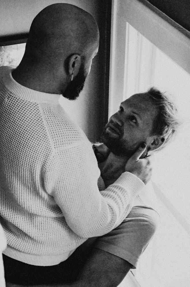 A man with a beard and hoop earring holding another man with dyed hair, in an intimate moment indoors.