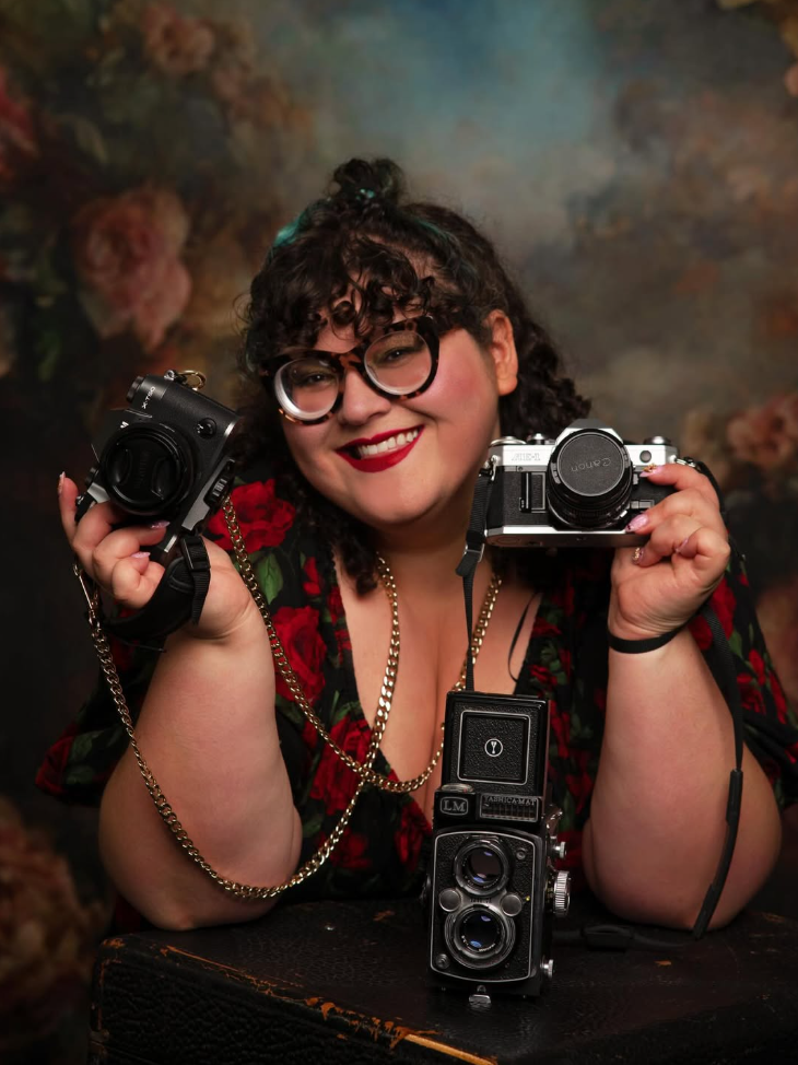 A woman with curly hair and glasses smiling, holding two cameras, with a vintage camera on the table in front of her, and a colorful background.