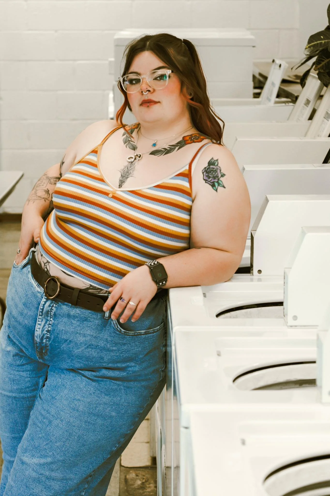 Photograph of a curvy woman with long dark hair, glasses, and tattoos, wearing a striped tank top and jeans, leaning against a white laundry machine in a laundromat.
