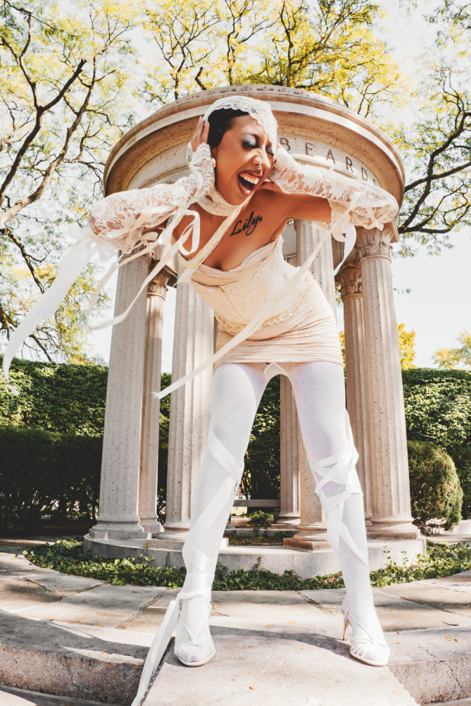 Creative photography of a woman dressing in a white outfit with white lace sleeves, white thigh-high stockings, and high heels, standing in front of a rounded stone structure with columns, with trees in the background.