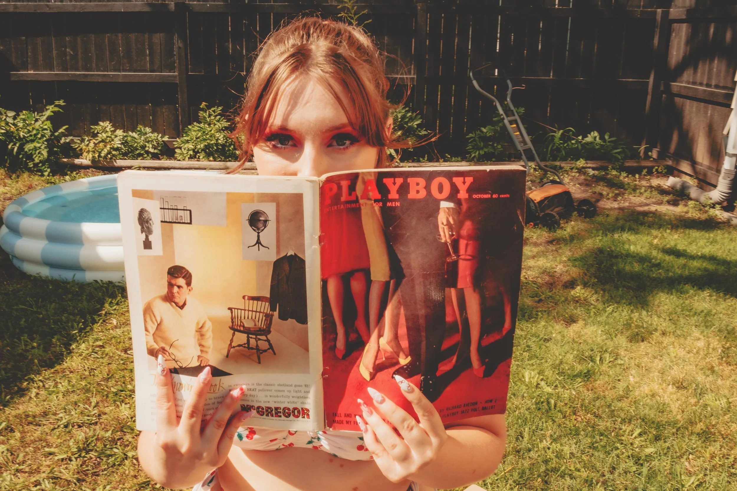 Person with red hair reading a vintage Playboy magazine outdoors in a backyard, with a small inflatable pool and a wooden fence in the background.