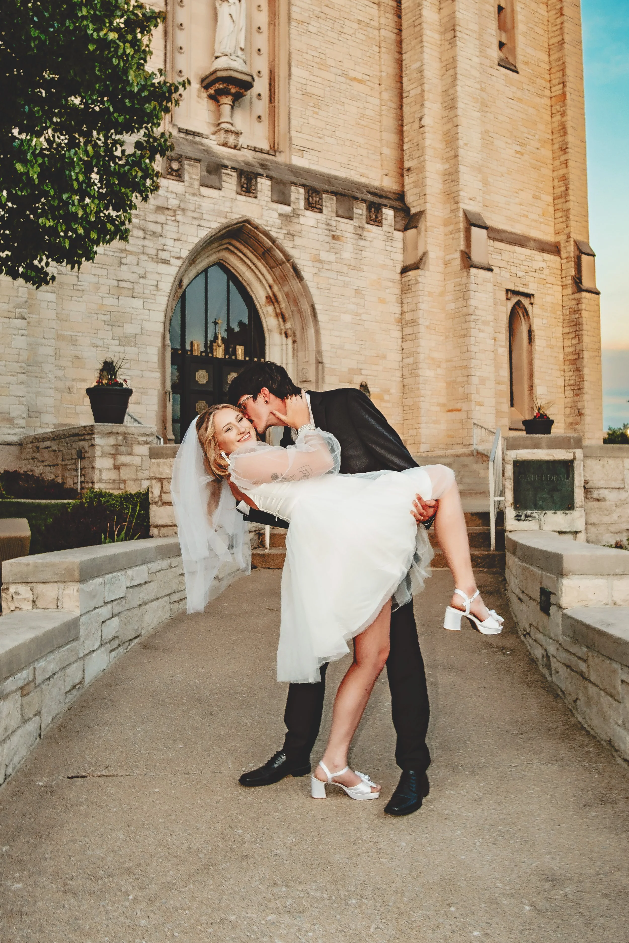 A couple dressed in wedding attire. The man in a black suit and the woman in a white dress, with the man lifting the woman in front of a stone church with gothic arches and a statue above the door.