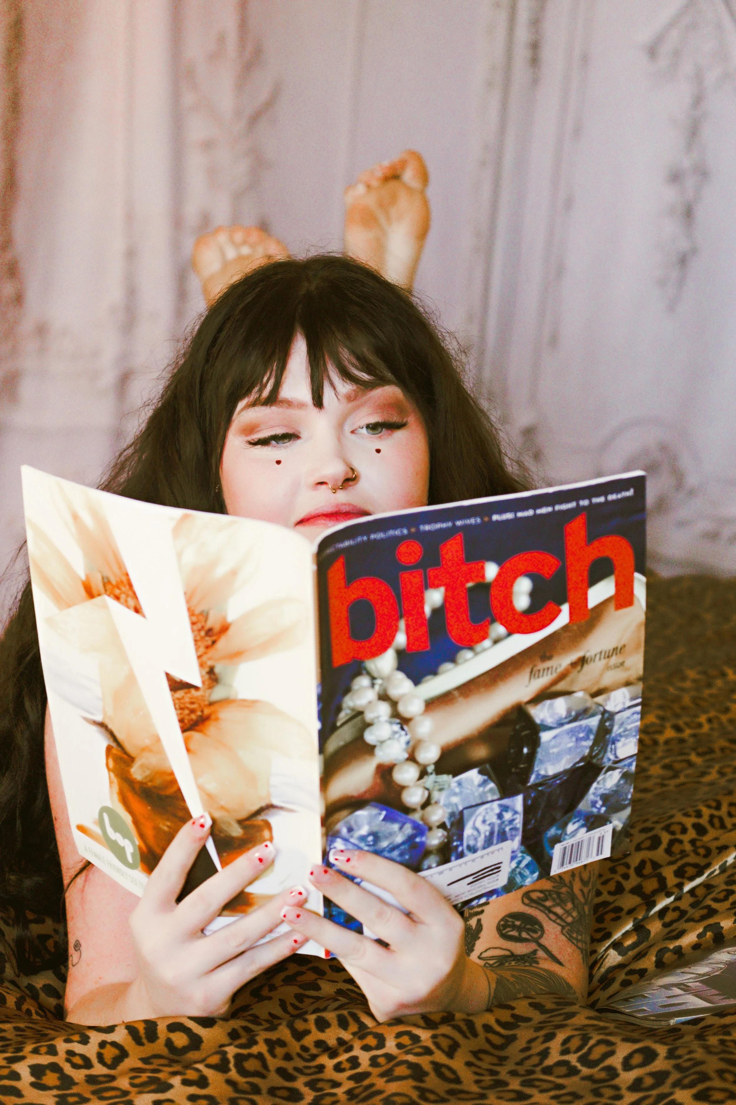 Woman with black hair, tattoos on arms, and makeup, reading a magazine with a woman on the cover, while lying on a leopard-print blanket.