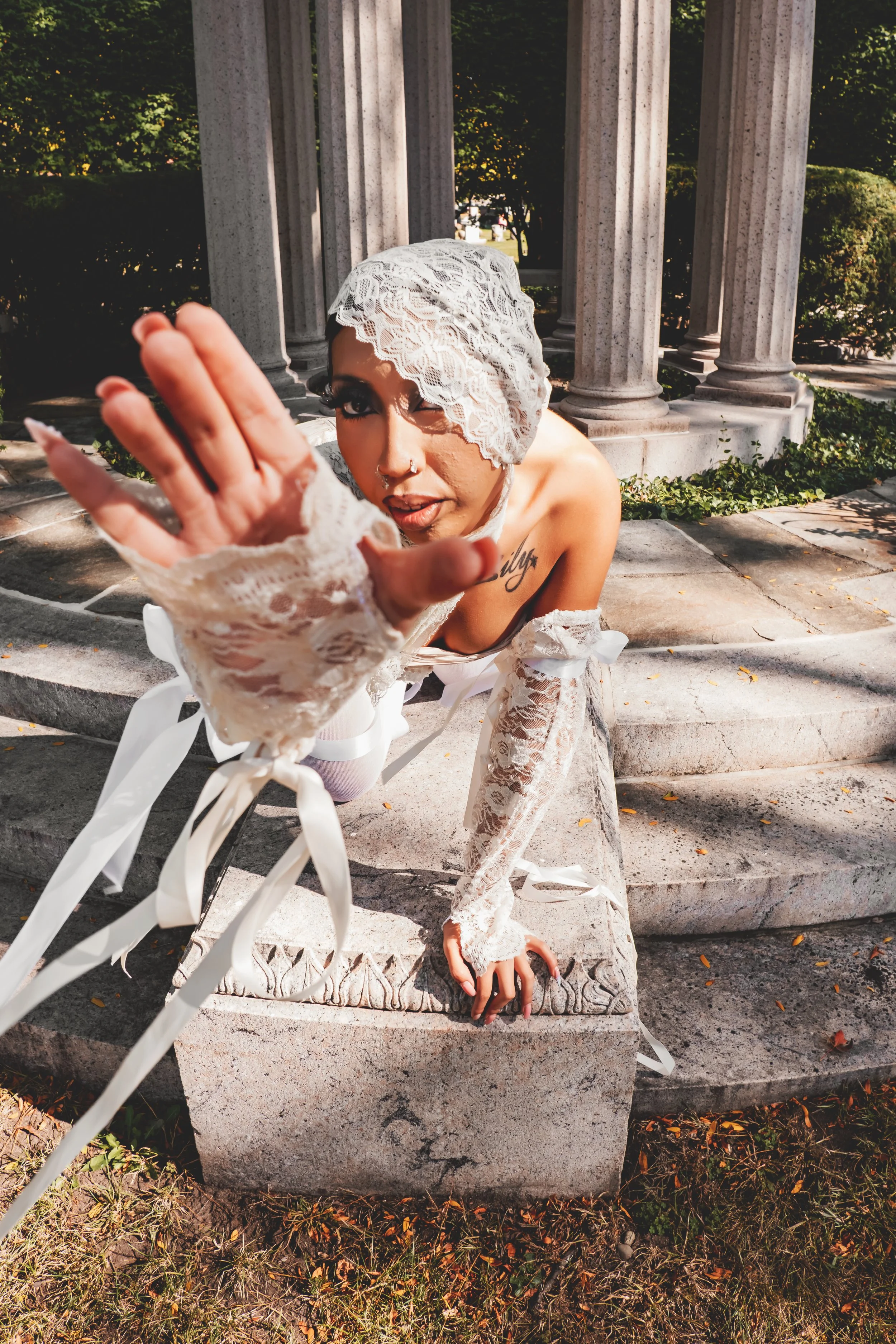 A woman with makeup and tattoos reaching towards the camera, wearing lace clothing and a lace head covering, leaning over a stone bench in an outdoor setting with trees and columns in the background.