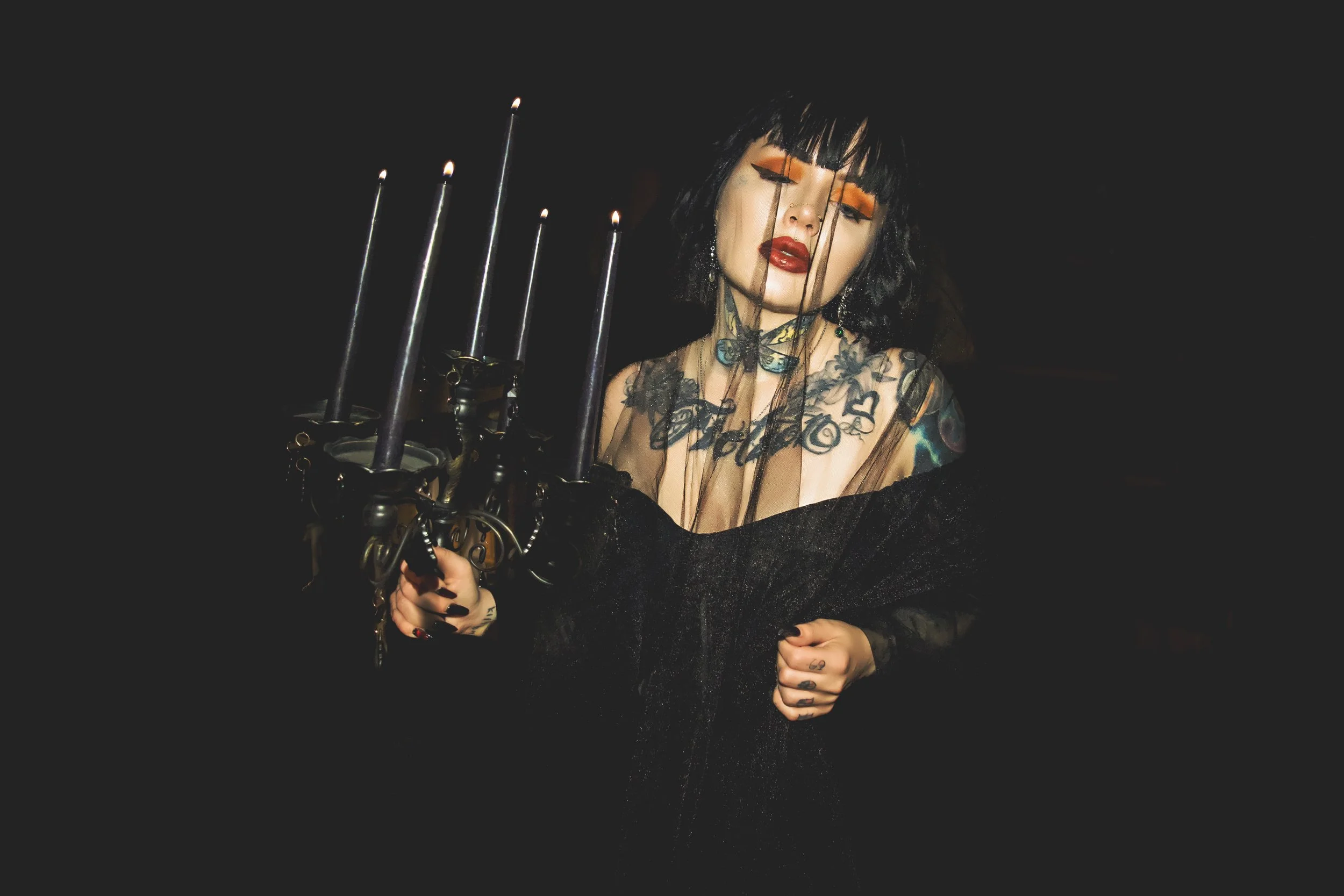 A woman with black hair and bangs, wearing orange eyeshadow and lipstick, holding a black candelabrum with lit candles, dressed in gothic-style clothing, with tattoos visible on her shoulders and arms, standing against a dark background.
