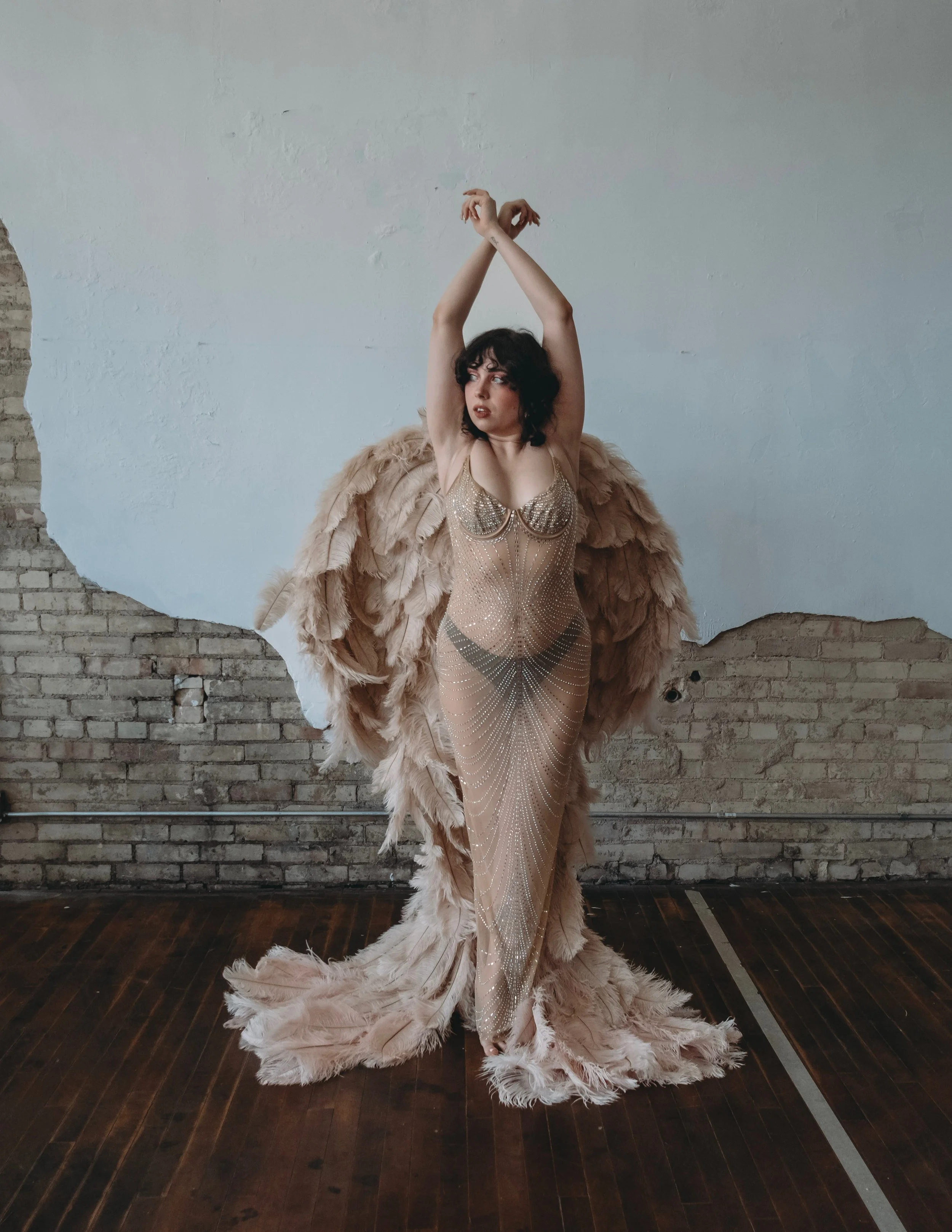 A woman wearing an ornate, beaded gown and large feathered wings stands against an unfinished brick wall, with arms raised above head, in an indoor setting.
