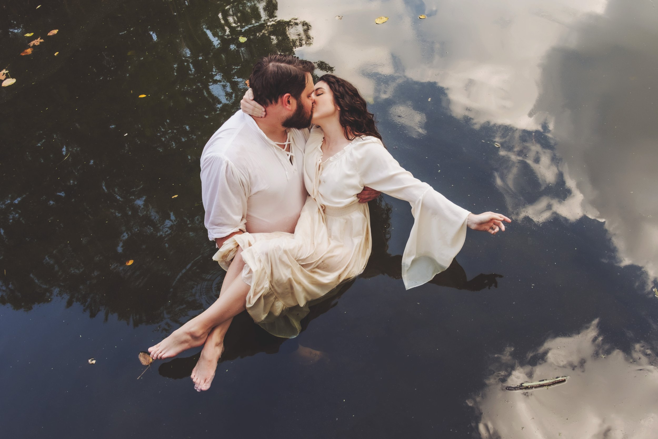 Photograph of a man and a woman sharing a kiss while floating in a body of water, with the woman in a cream-colored dress and the man in a white shirt. The water reflects the sky and trees, and there are a few scattered leaves on the surface.