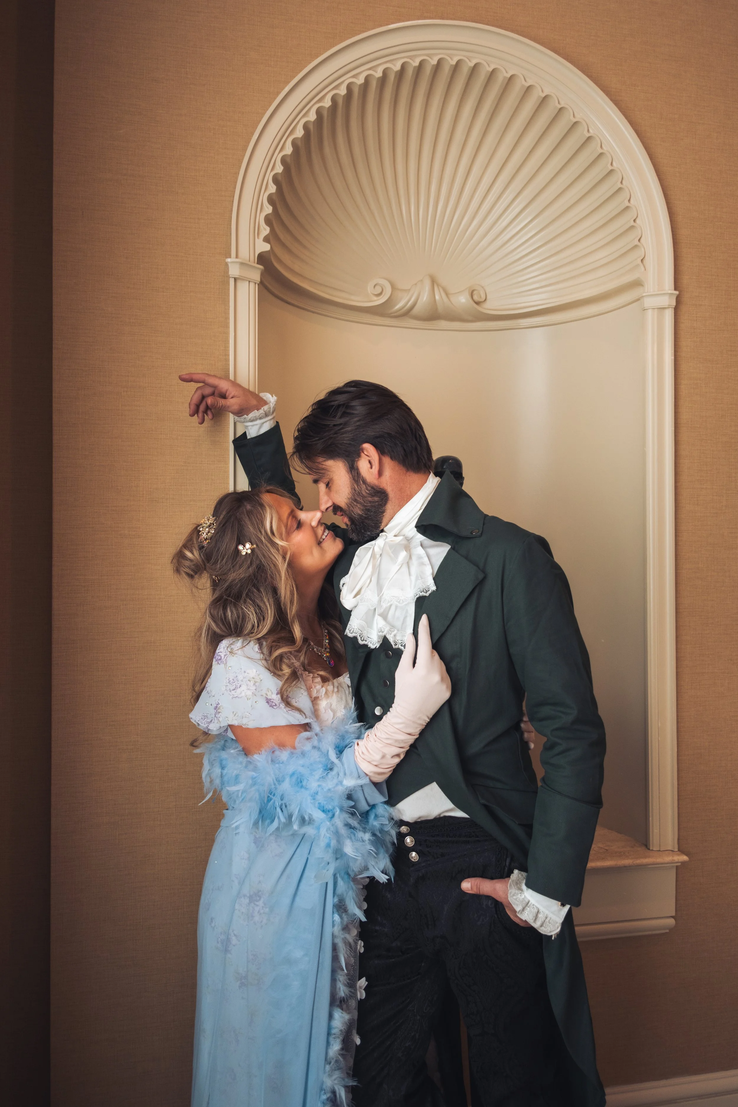 A man and woman in historical costume sharing an intimate moment on a dance floor against a beige wall with a decorative shell-shaped relief.
