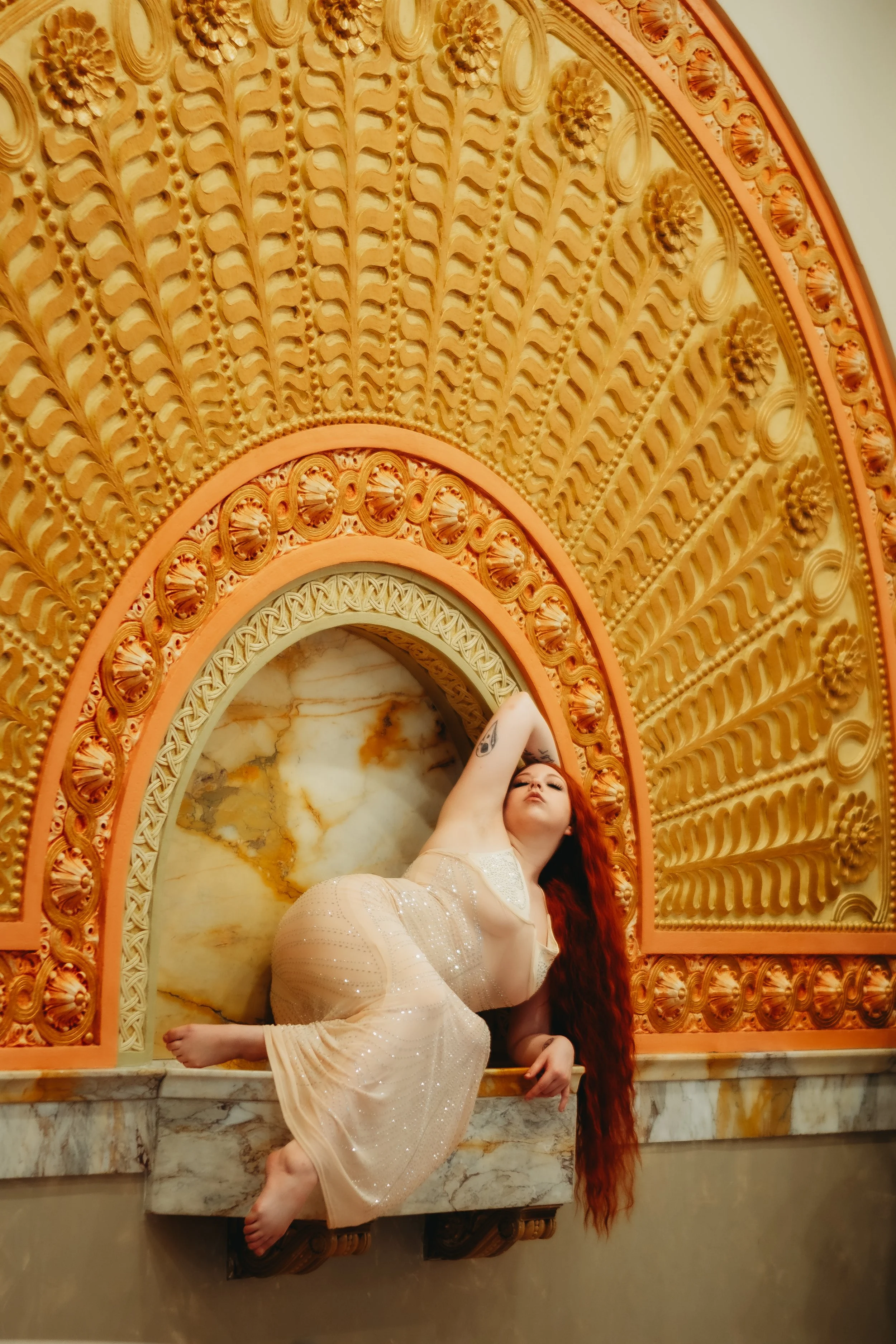 A woman with long red hair and tattoos on her arm, wearing a sheer, shimmering beige dress, posing gracefully against an ornate, circular gold and marble decorative background.