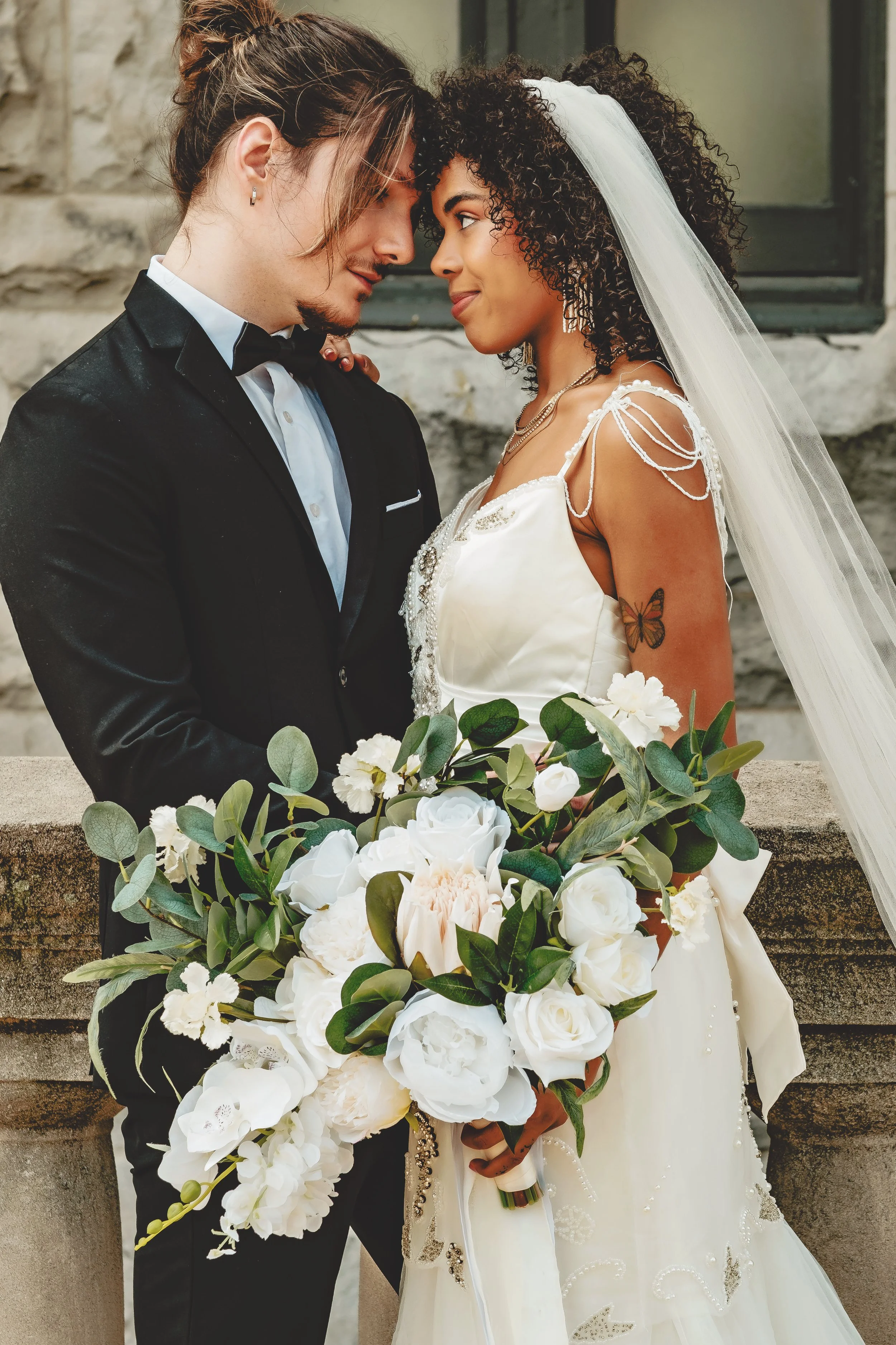 A couple on their wedding day, standing close with foreheads touching, dressed in formal wedding attire, with a bouquet of white flowers in the foreground, against a stone wall background.