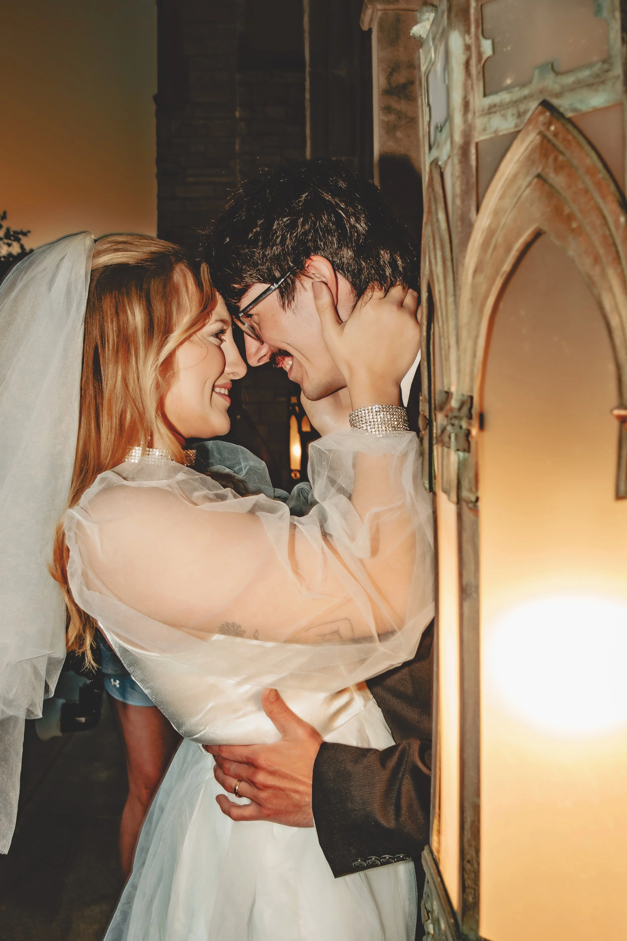A bride and groom are close together, touching foreheads and smiling at each other at their wedding. The bride has long red hair and wears a veil, while the groom has dark hair and wears glasses and a suit. They are in a warmly lit room with a vanity