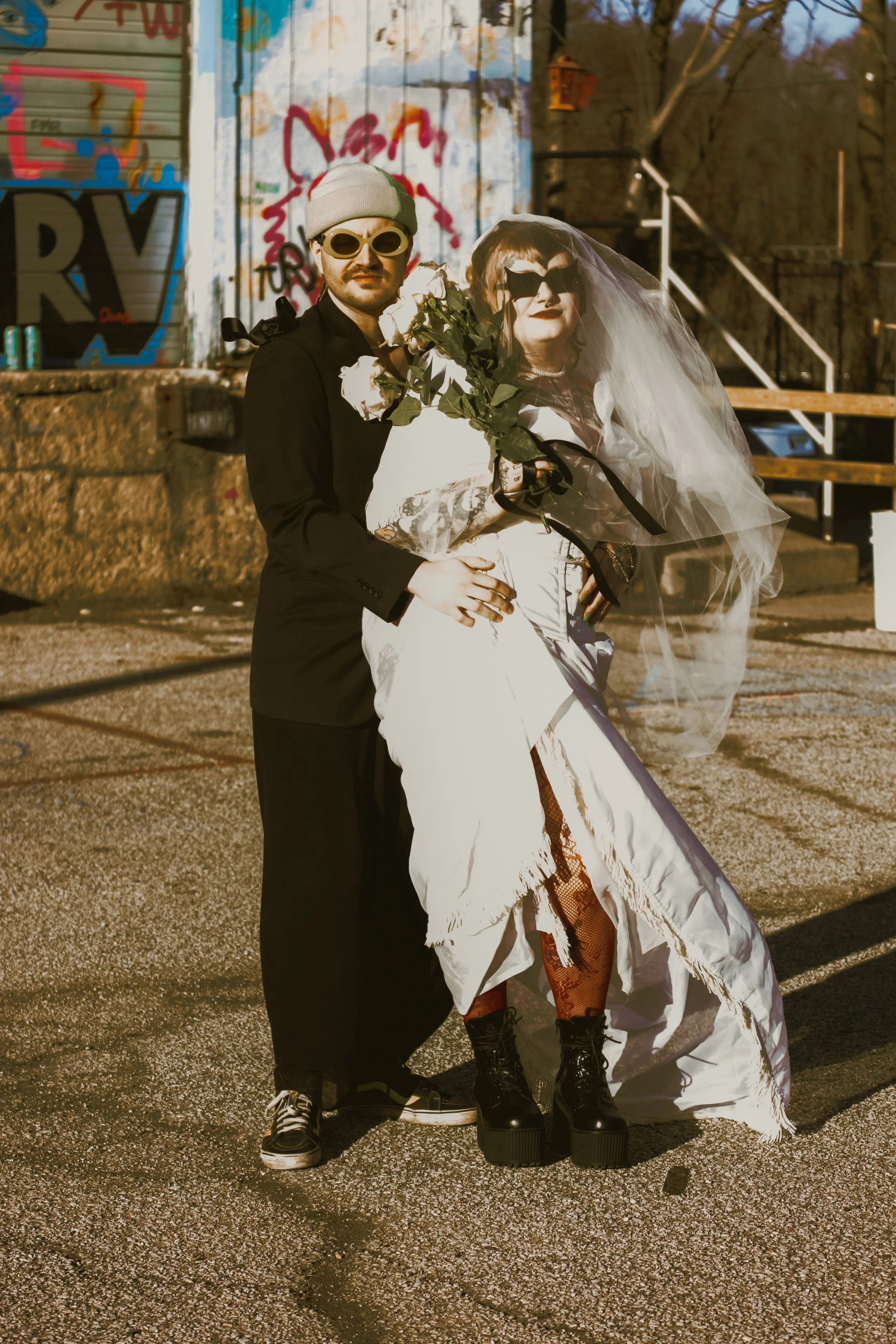 Two individuals dressed in alternative fashion, standing outdoors near graffiti-covered walls, with one holding a bouquet of white roses, both wearing sunglasses, one with a veil, and the other with a beanie.