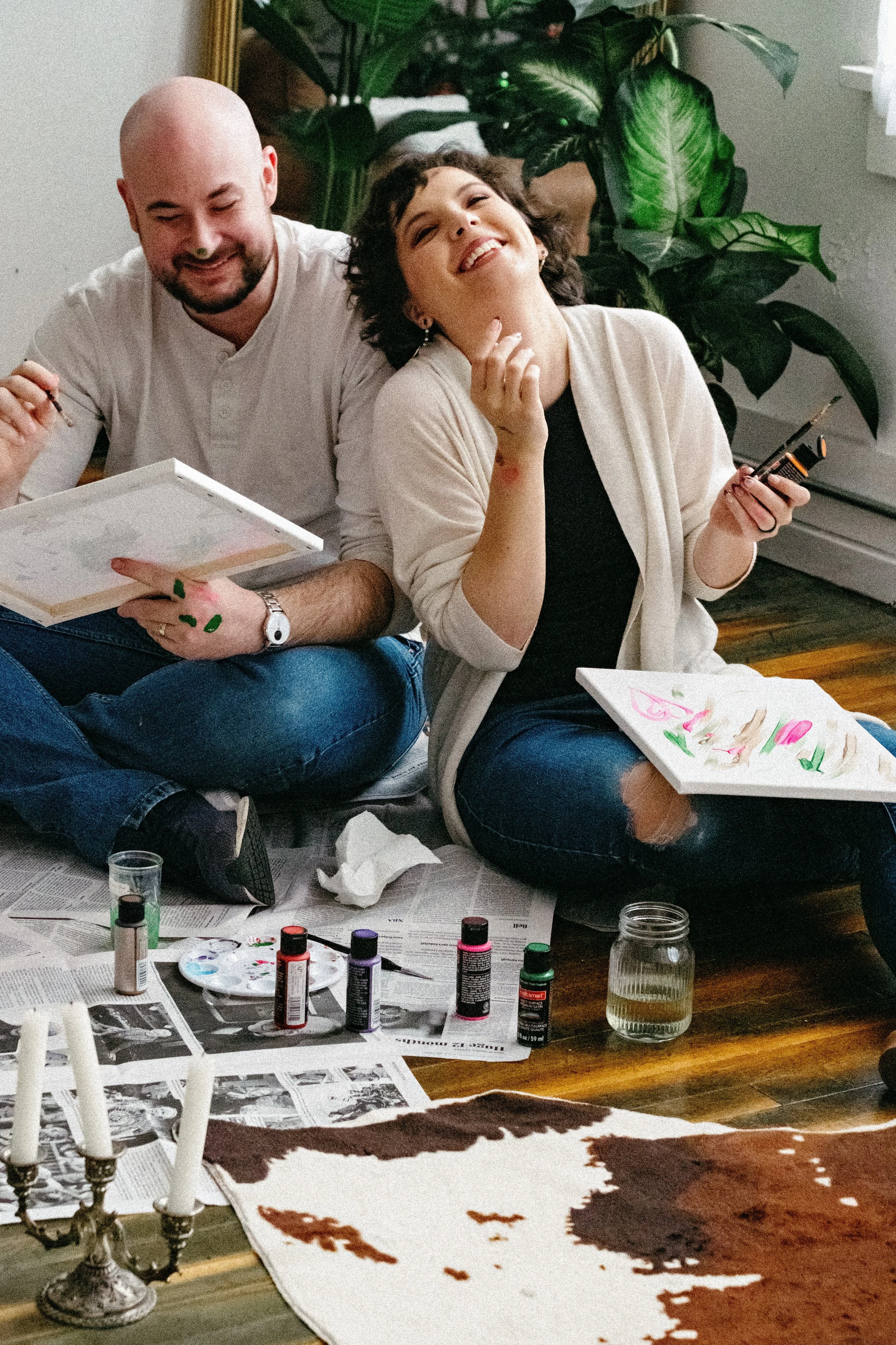 Two people sitting on the floor in a room, engaged in painting or craft activity. The man, on the left, has a beard and is holding a paintbrush and a palette. The woman, on the right, has curly hair and is smiling, holding a paintbrush and palette, w