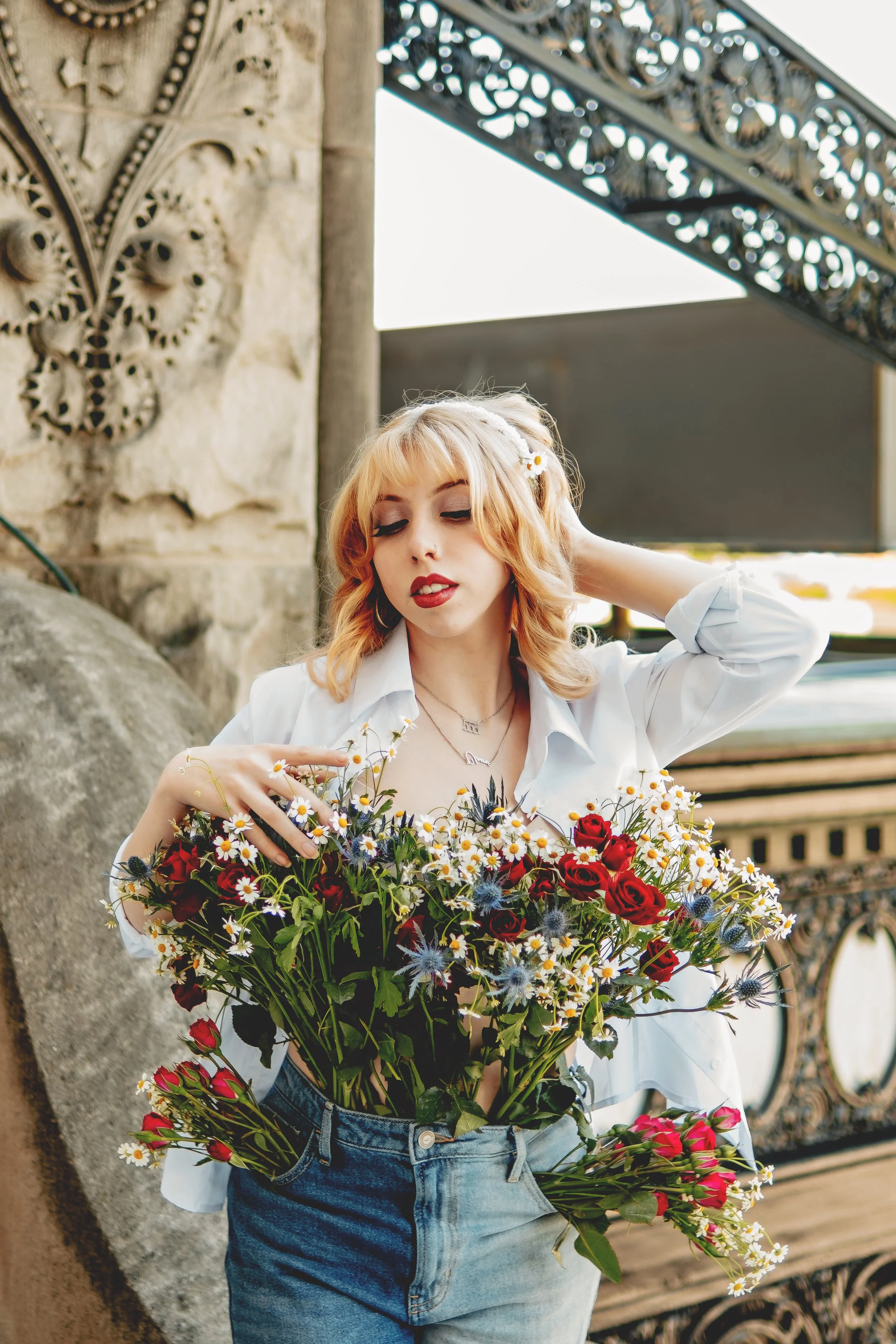 A young woman with blonde hair and red lipstick holding a large bouquet of flowers, including daisies and roses, standing outdoors near a stone wall and decorative metalwork.