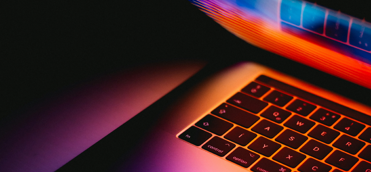 Close-up of a laptop keyboard illuminated with orange and purple backlighting, with part of the laptop screen visible in the background.