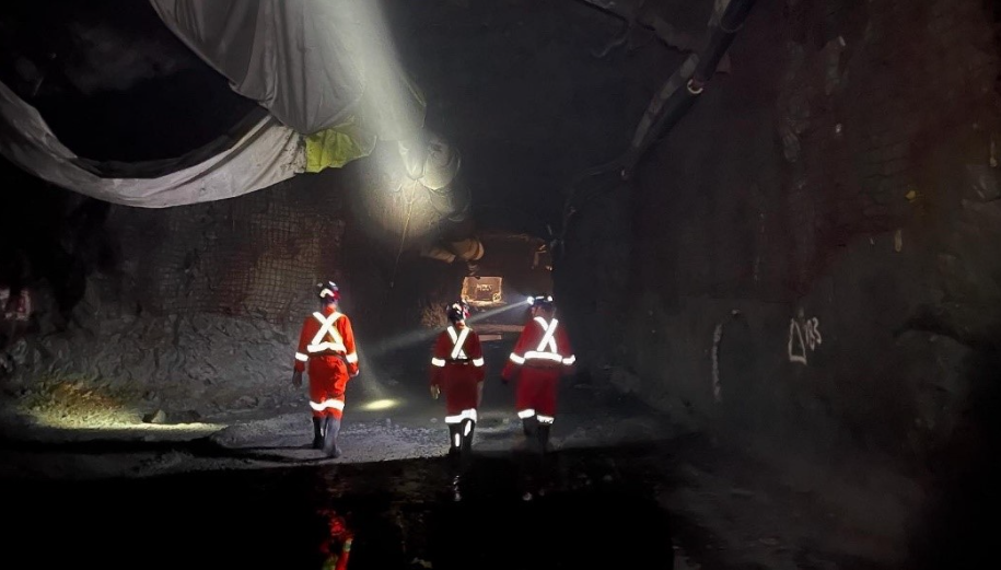 Three workers in reflective safety gear walking through a dark underground tunnel or mine shaft, with construction equipment visible in the distance.