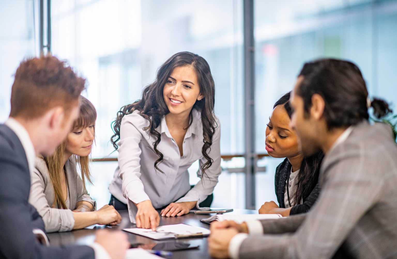A woman standing and gesturing at a table with five seated colleagues in a business meeting.