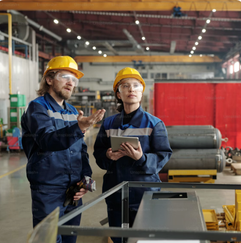Two factory workers wearing yellow safety helmets and glasses, standing and discussing in an industrial setting with machinery and equipment in the background.