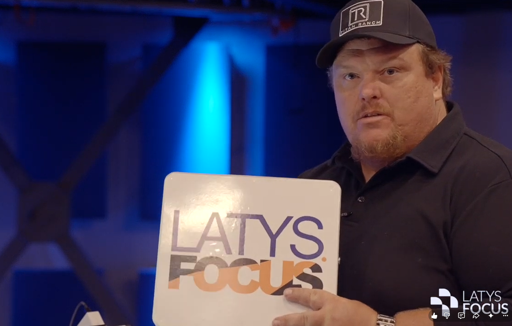 A man wearing a black cap and black shirt holds a book titled "LATYS FOCUS" in front of him at an indoor event with blue lighting.