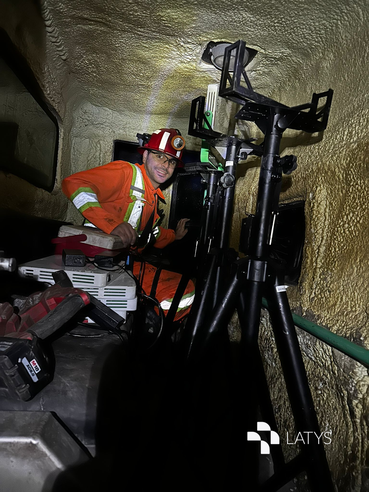 A male worker wearing an orange safety suit and a red helmet with a headlamp inside a mining tunnel, surrounded by mining equipment and tools.