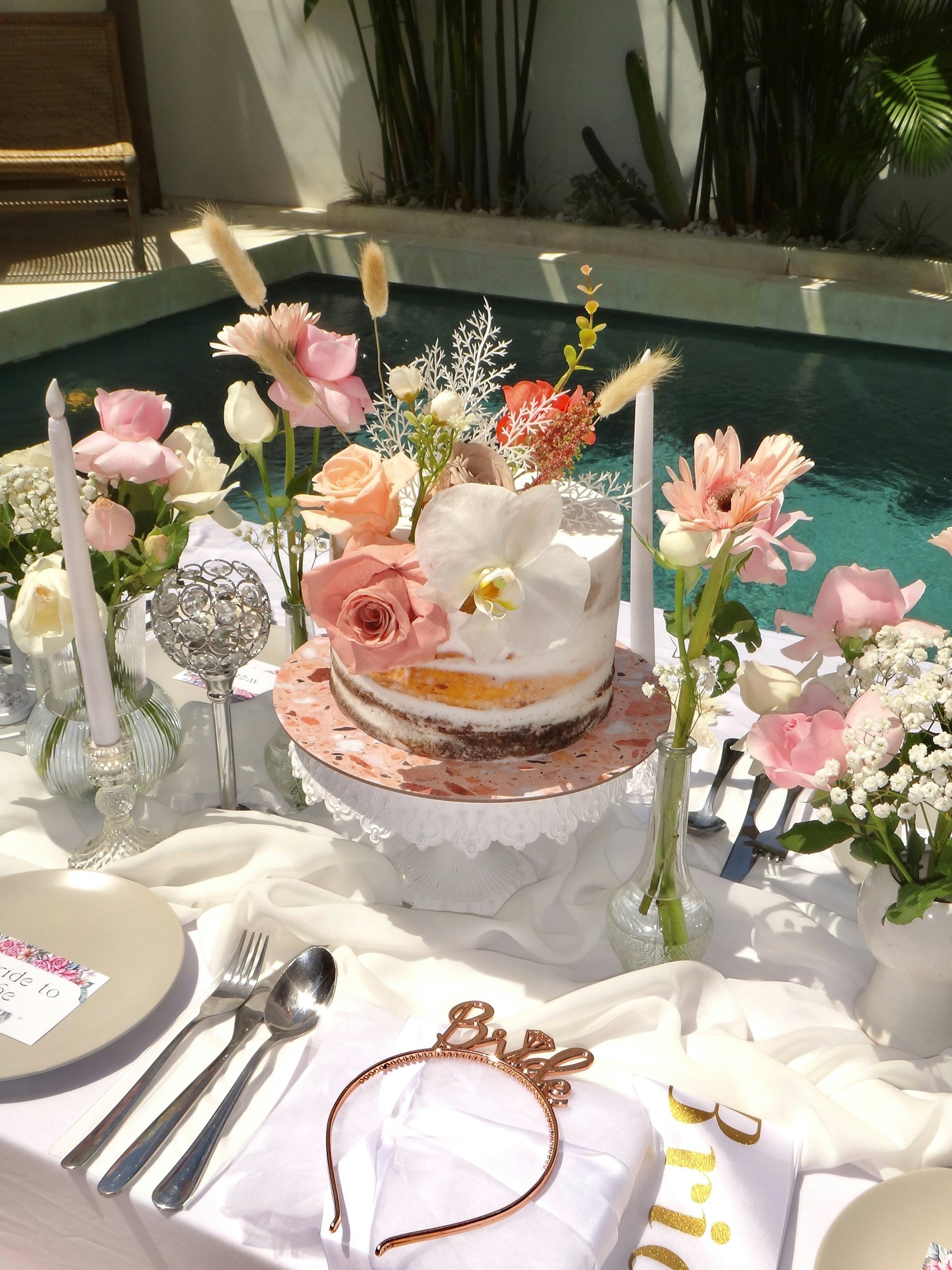 Decorative table setting with a cake topped with flowers, surrounded by vases with flowers, candles, and utensils, on a white tablecloth, outdoors with a pool and greenery in the background.