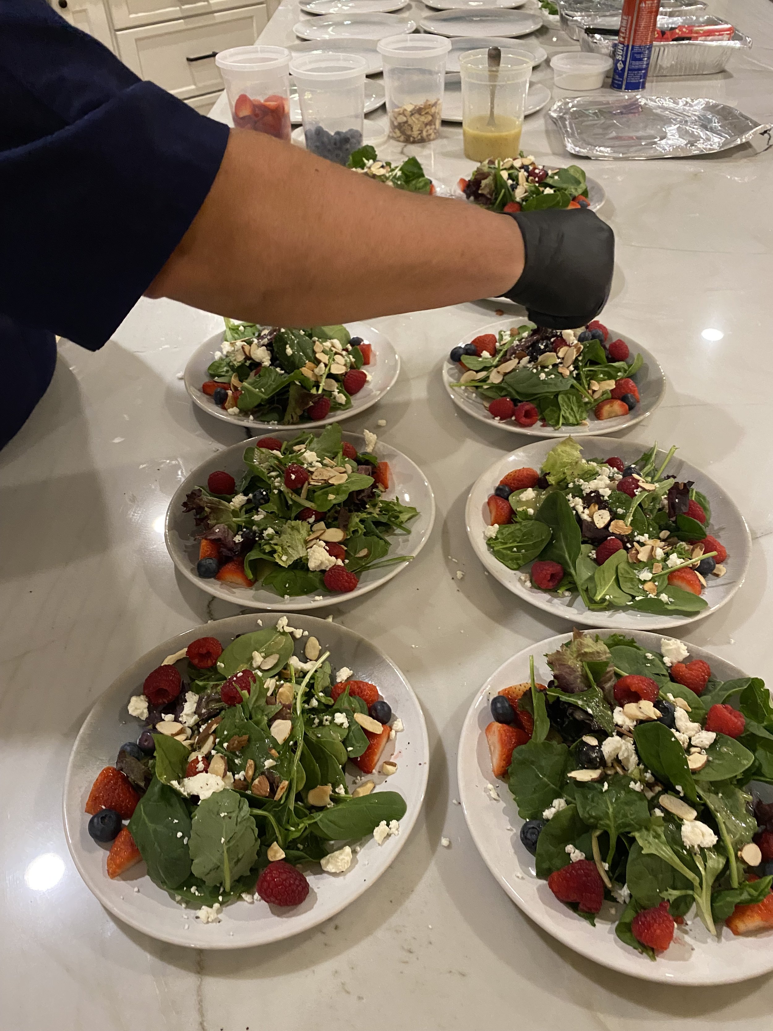 Multiple plates of fresh green salad with strawberries, blueberries, raspberries, nuts, and cheese on a white table, with condiments and ingredients in small containers in the background.