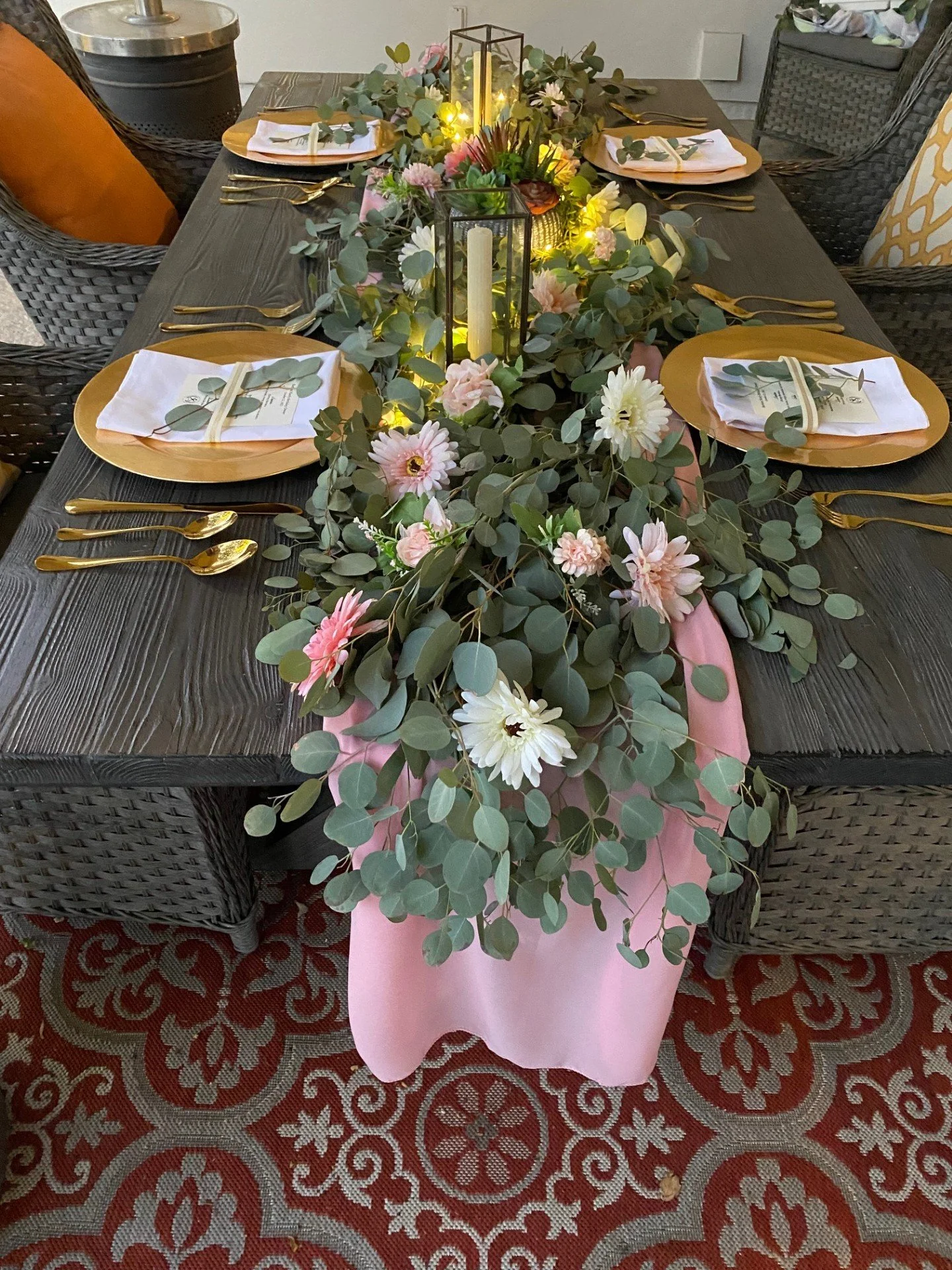 A beautifully decorated table with a floral centerpiece with pink and white flowers, greenery, and candles.  The table setting includes gold cutlery, white napkins, and plates with a custom menu for a luxury outdoor dinner party in Scottsdale.