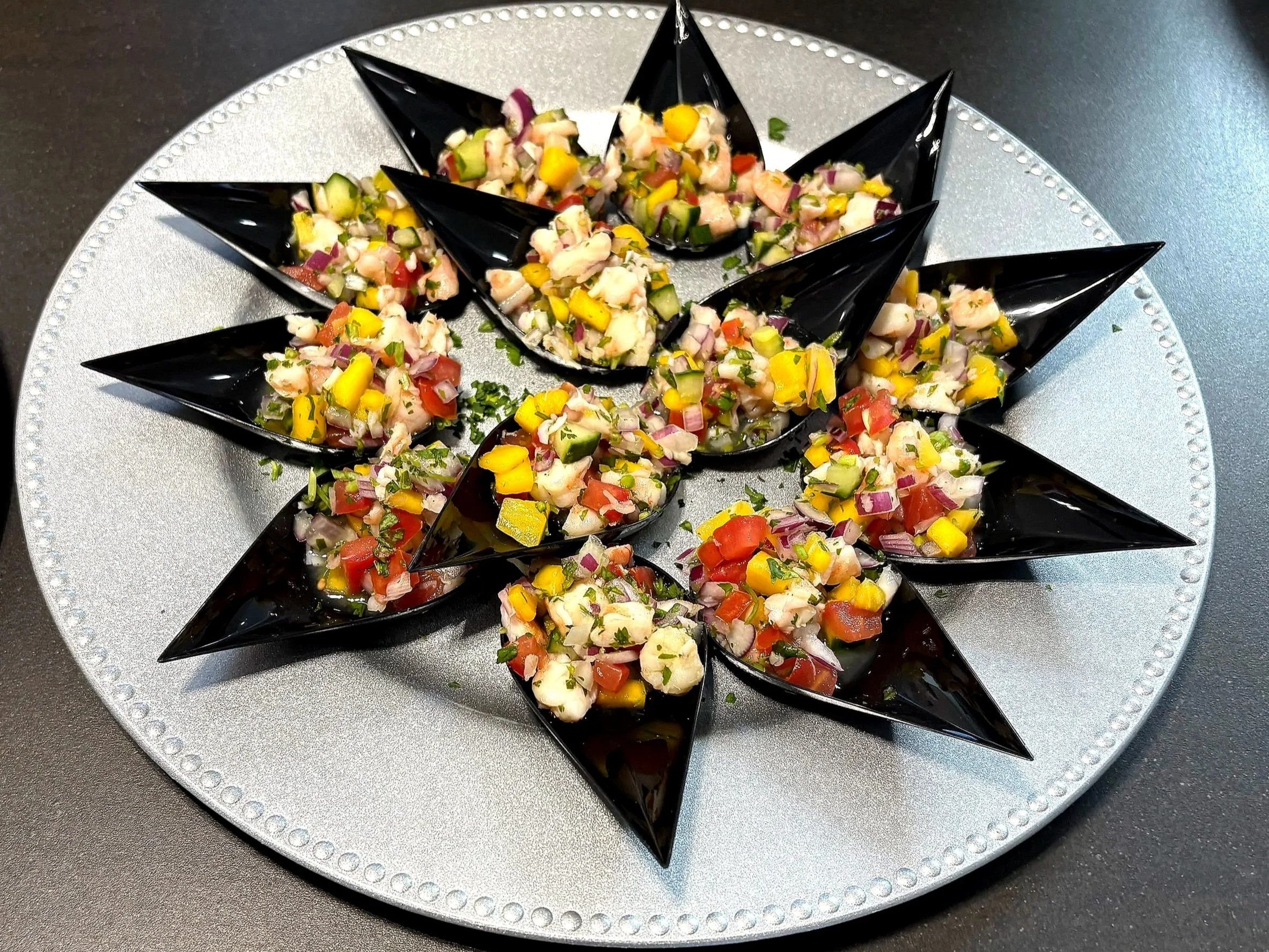 Plate of shrimp ceviche topped with chopped vegetables on black spoon-shaped containers arranged in a circular pattern on a silver platter.