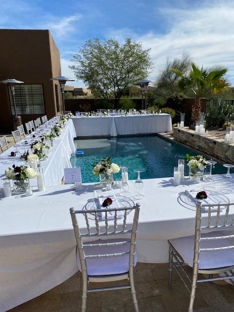 Wedding reception setup around a backyard swimming pool with long white tables, floral centerpieces, candles, and white chairs, under a partly cloudy sky with desert plants and trees in the background with a luxury plated dinner service in Carefree.