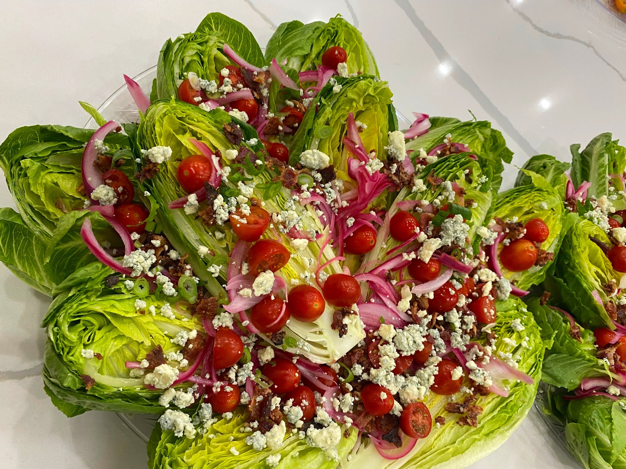 Wedge Salad with lettuce, cherry tomatoes, red onions, blue cheese crumbles, bacon bits, and green onions in a glass bowl on a white surface.