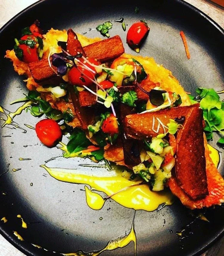 A plated dish with crispy tofu, cherry tomatoes, mixed greens, with a drizzle of yellow sauce and olive oil on a black plate.