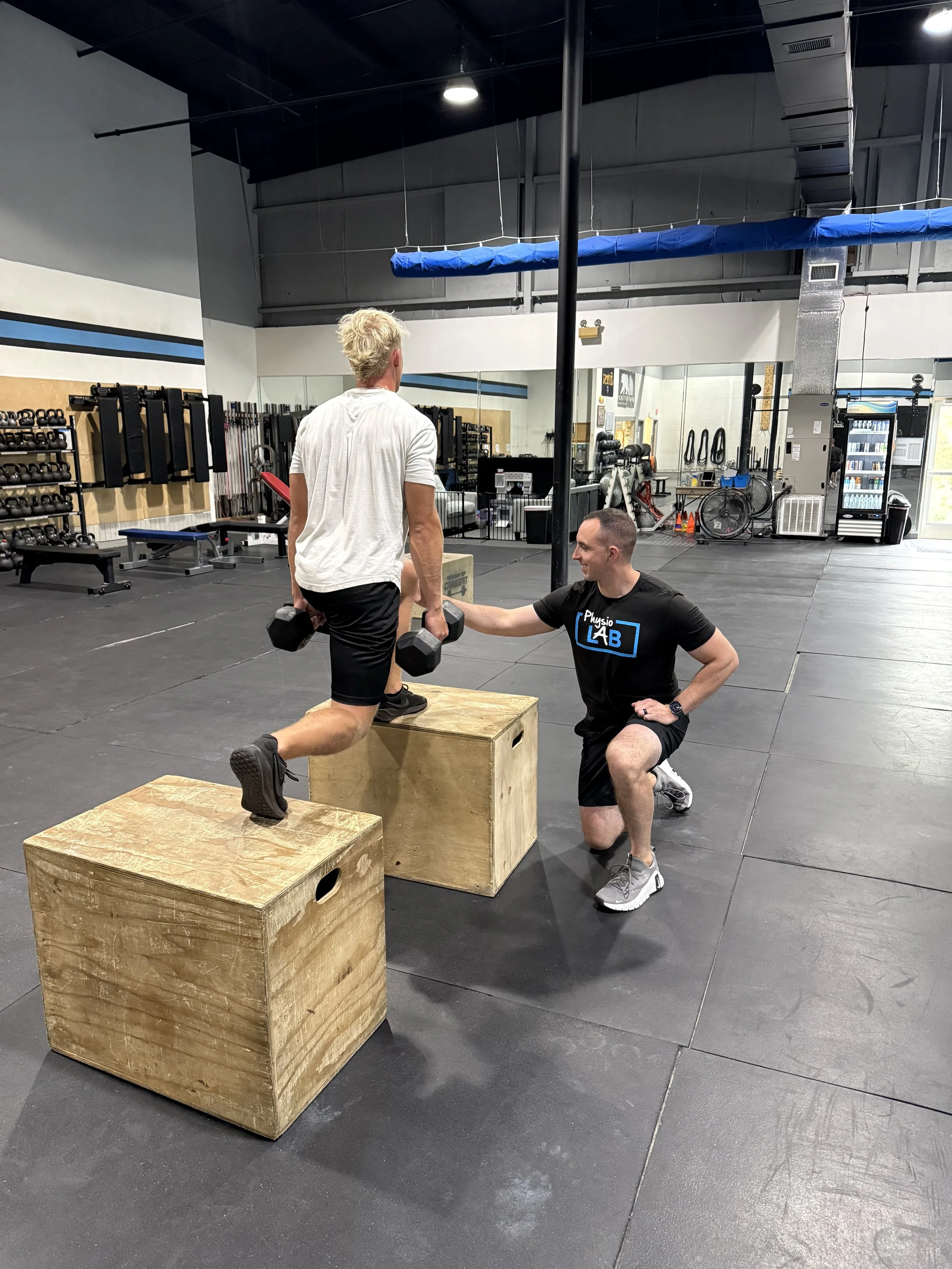 A man performs a lunge exercise with dumbbells while a trainer offers guidance at a gym. The gym has black flooring, wooden plyometric boxes, and fitness equipment including kettlebells, barbells, and a vending machine.