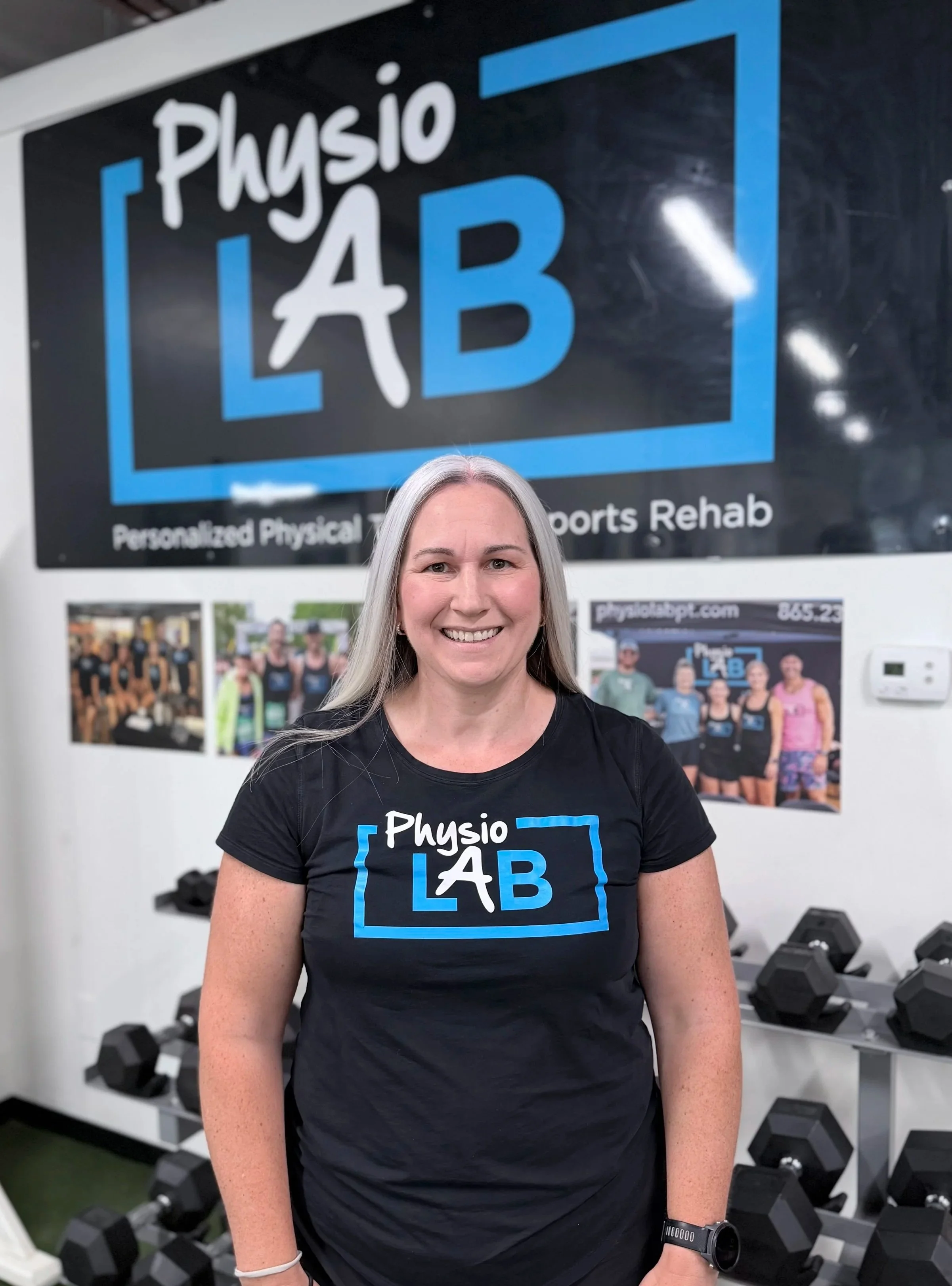 A woman with long gray hair smiling, standing in a gym, wearing a black t-shirt with the logo 'Physio LAB' on the front, and a black watch on her left wrist. Behind her are black dumbbells and a wall with fitness posters.