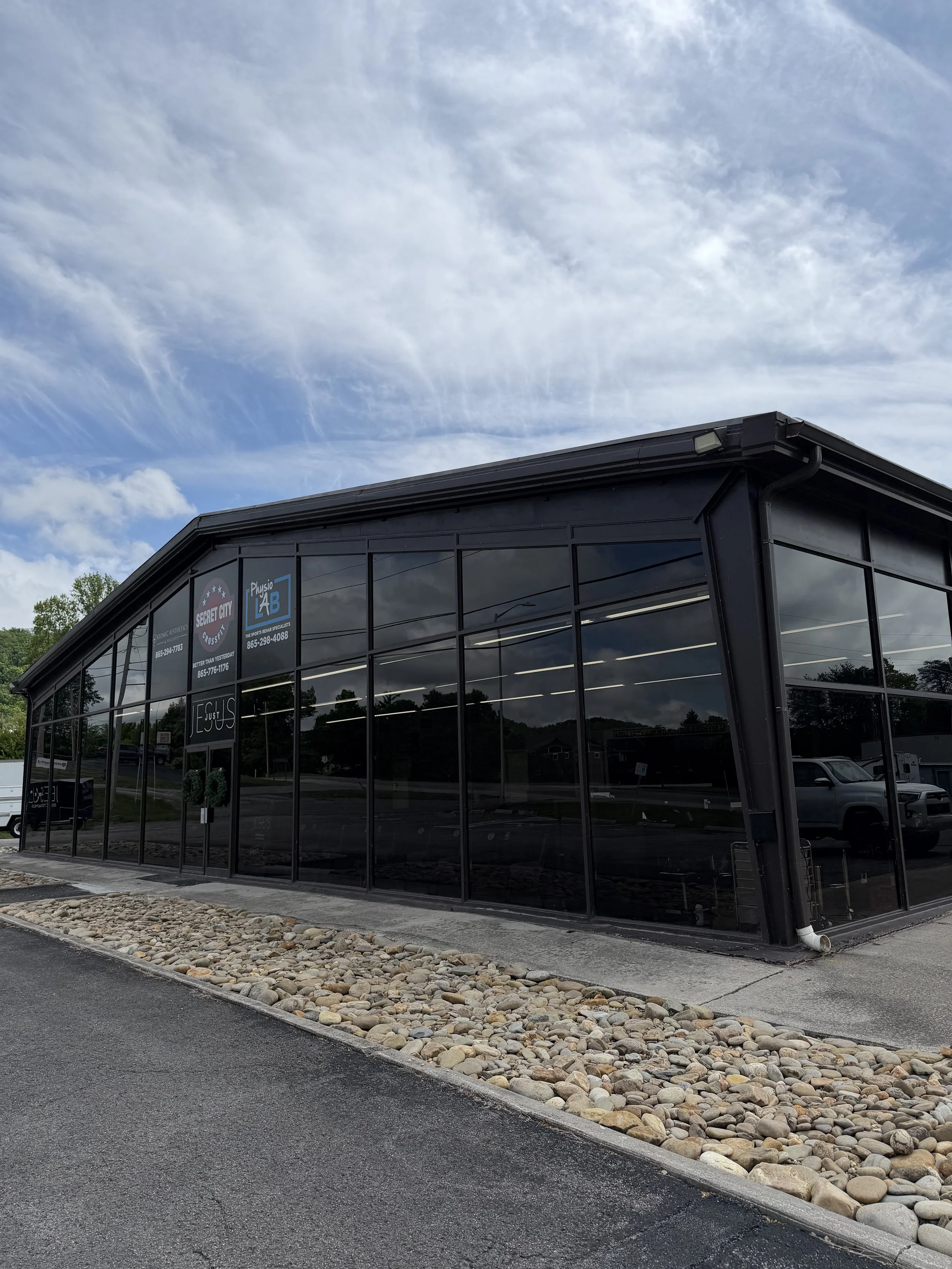 A modern building with a black exterior, large glass windows, and signs for fitness businesses, located next to a parking lot with a decorative rock border and a cloudy sky overhead.