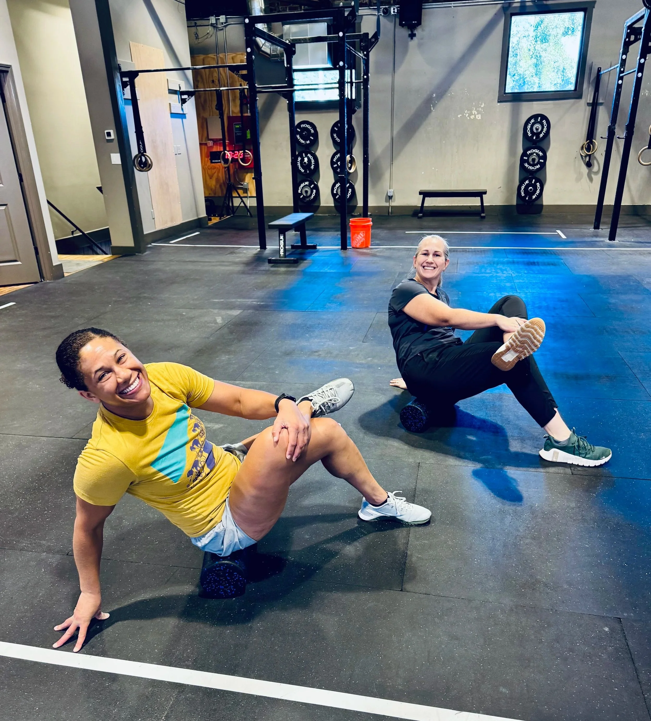 Two women resting on the floor of a gym, smiling. The gym has black flooring, exercise equipment, a window, and a wall mounted digital clock showing 07:23.