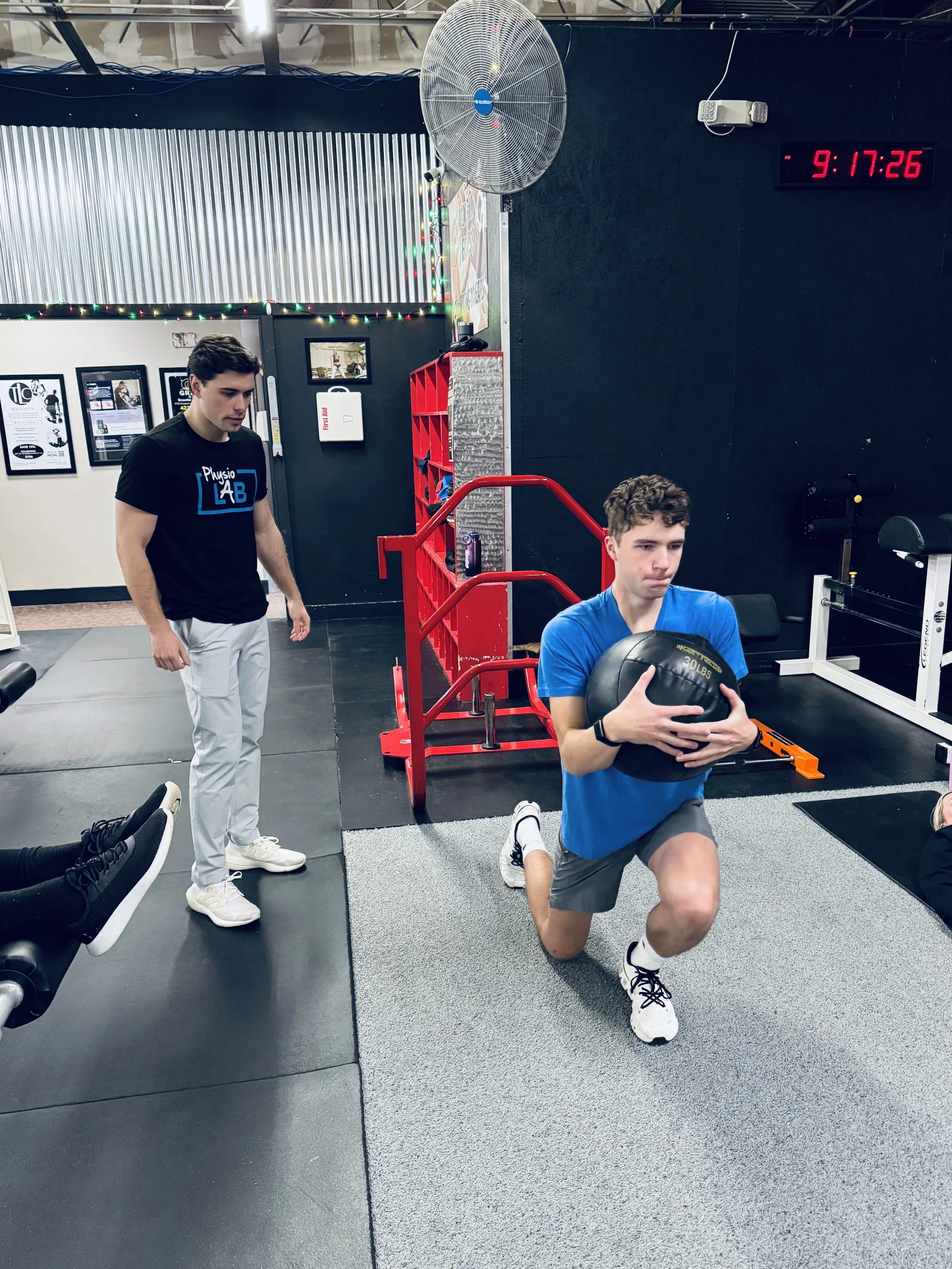 A young man is performing a lunge exercise with a medicine ball in a gym. He is wearing a blue shirt, gray shorts, and white sneakers. Another man is standing nearby, observing and wearing a black T-shirt and white pants. The gym has black flooring, a gray mat, and fitness equipment with a black wall in the background. The wall displays a digital clock showing 9:17:26, and there are colorful lights and posters on the walls.