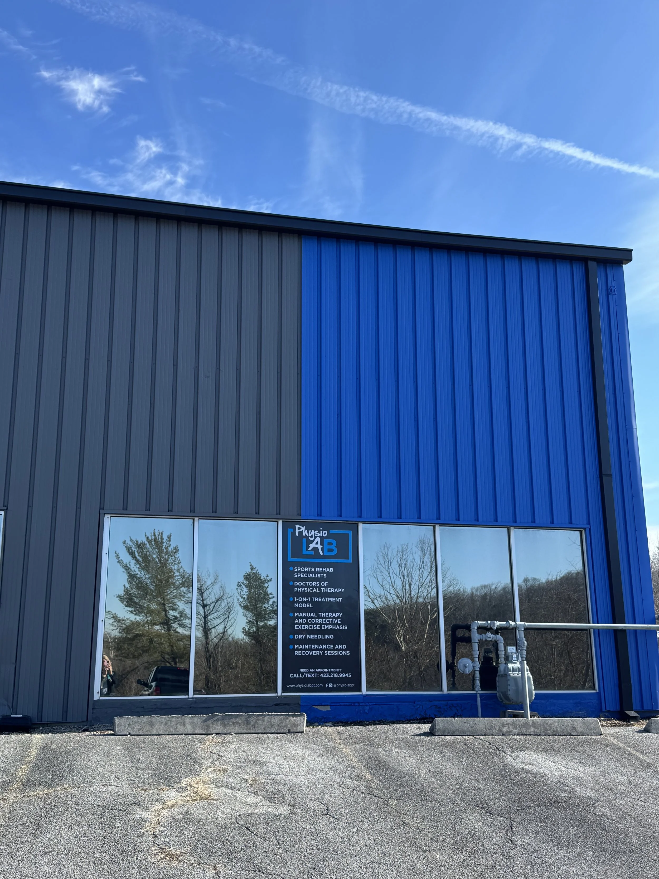 Image of a building with a large window and a sign for Physio Lab, a sports rehab and physical therapy clinic. The sign details services such as sports rehab, manual therapy, dry needling, and recovery sessions. Outside, there is a parking lot with cracks and concrete barriers, and some trees are reflected in the window.
