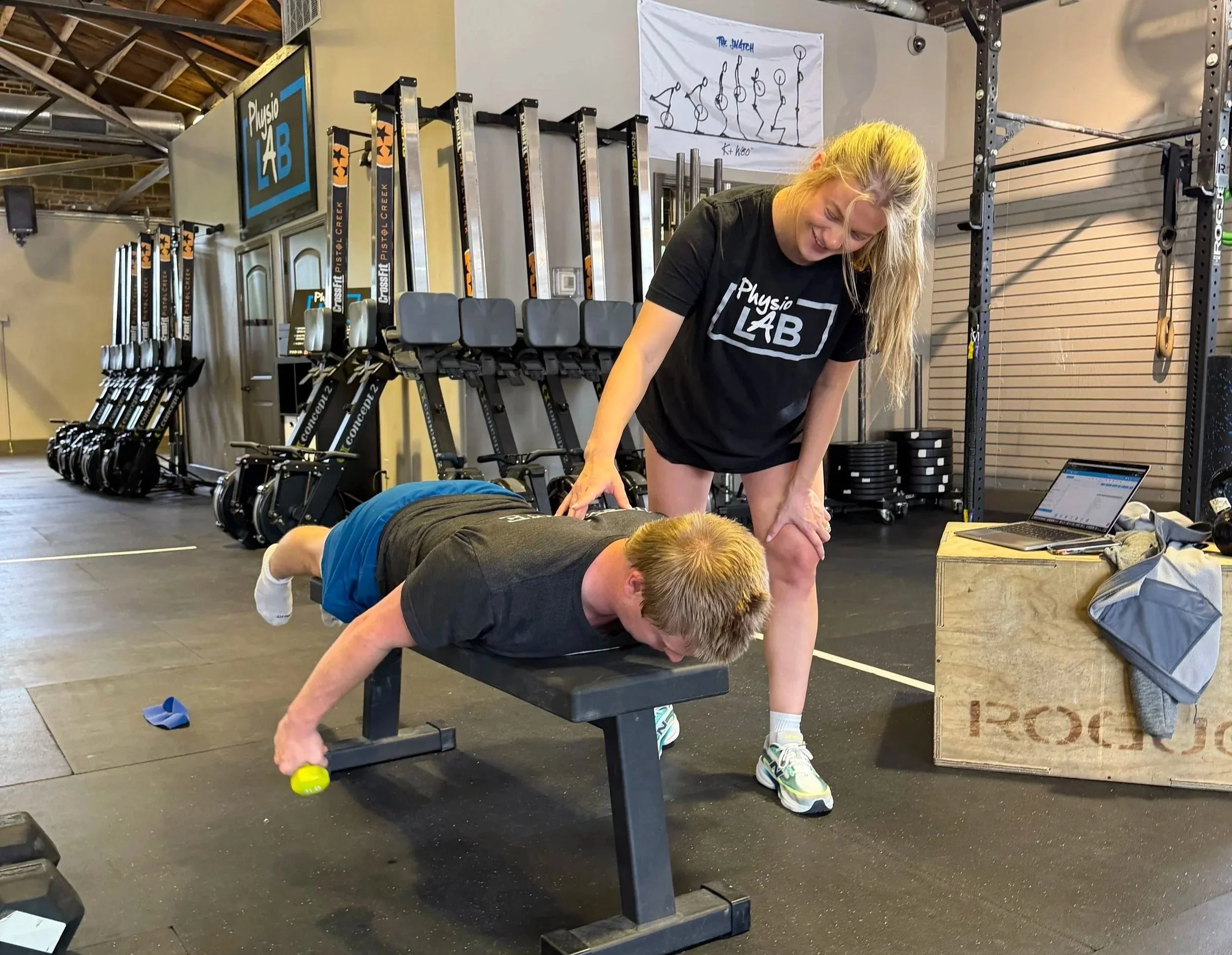 A woman is performing a push-up exercise on a black workout bench in a gym. She is smiling and wearing a black shirt with the words 'Physio LAB' on it. Behind her, a man is on the floor in a plank position, holding a yellow object in his hand. The gym has various equipment, including rowing machines, black weight plates, and a wall-mounted row of kettlebells. A sketch of yoga poses is visible on a whiteboard, and some equipment is stored on racks.